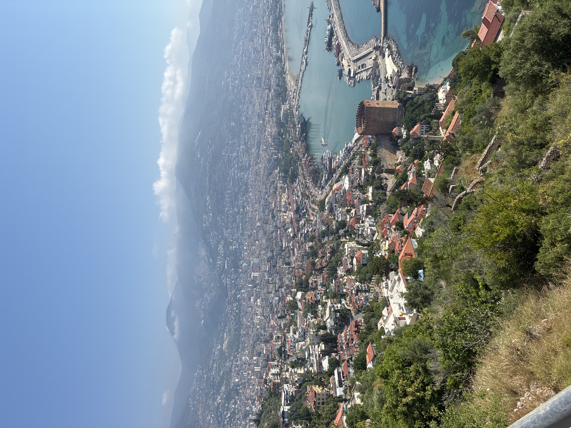 This is a high-angle, landscape photograph of Alanya, Turkey, taken on a clear, sunny day with a few clouds in the distant sky. The foreground features a hillside covered in green vegetation and dry grass, with a metal railing visible on the left.
The midground showcases the city of Alanya, characterized by its densely packed buildings with terracotta roofs, ascending the hillsides. Prominent features include a large, circular red brick tower on a cliff overlooking the sea, likely part of the Alanya Castle complex. Below the tower, a bay is visible, filled with numerous boats of varying sizes, including several large sailboats docked along the harbor. A breakwater extends into the turquoise water, with more boats moored along its length. A small beach with a few people is visible near the base of the castle.
The background reveals a sprawling urban landscape stretching along the coastline, with more buildings climbing the steep, hazy mountains that form the backdrop. The overall impression is of a vibrant coastal city nestled between mountains and the sea.