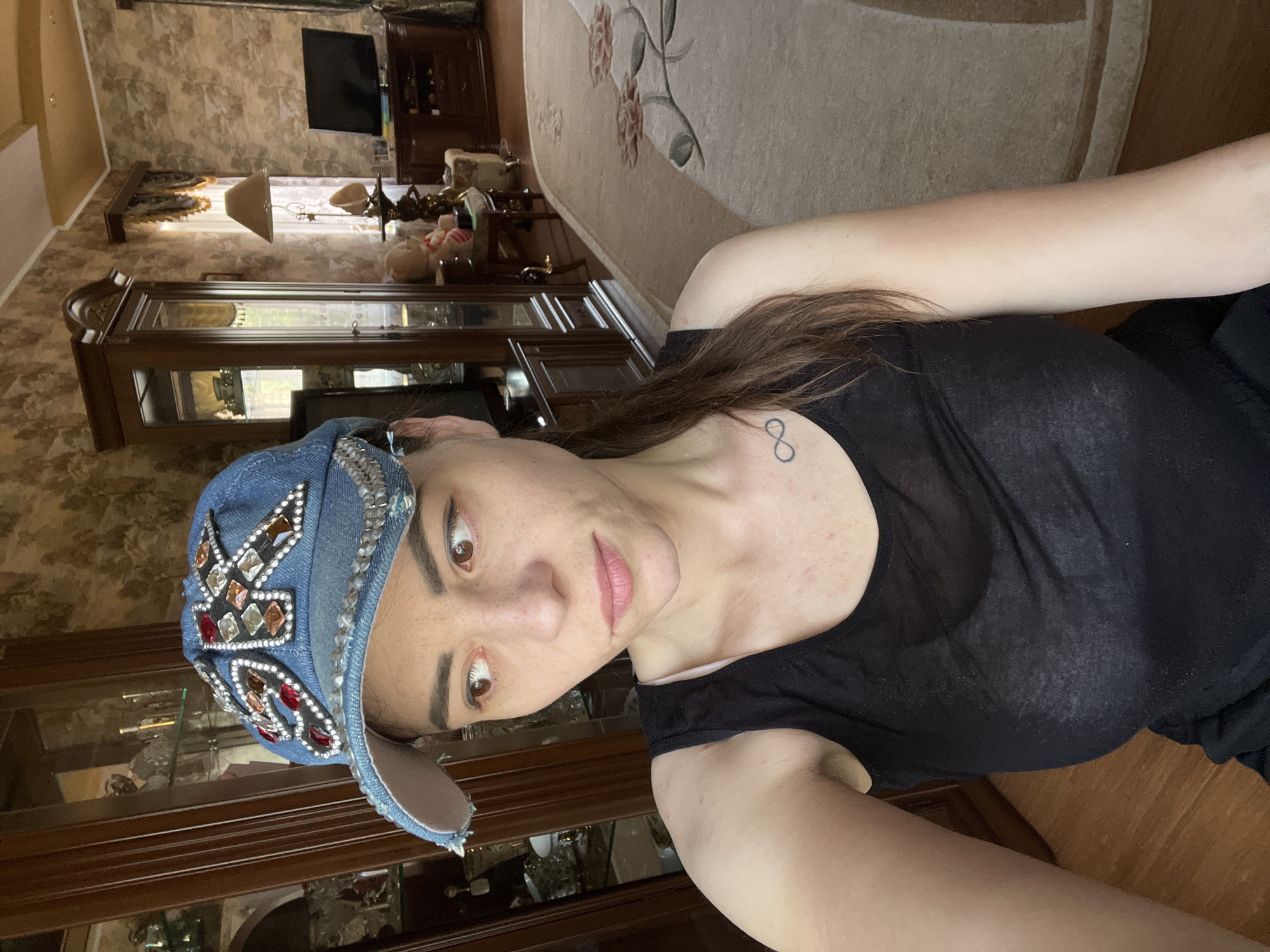 A young woman with brown hair, wearing a black tank top and a denim cap adorned with rhinestones, poses for a selfie in a room with traditional Russian decor. The room features dark wood furniture, including a display cabinet filled with glass objects, a large wooden hutch, and a plush rug with floral patterns. A television is visible in the background, along with a floor lamp and a table with a stuffed toy. The woman has an infinity tattoo on her collarbone. The scene appears to be indoors, likely in a residential setting.