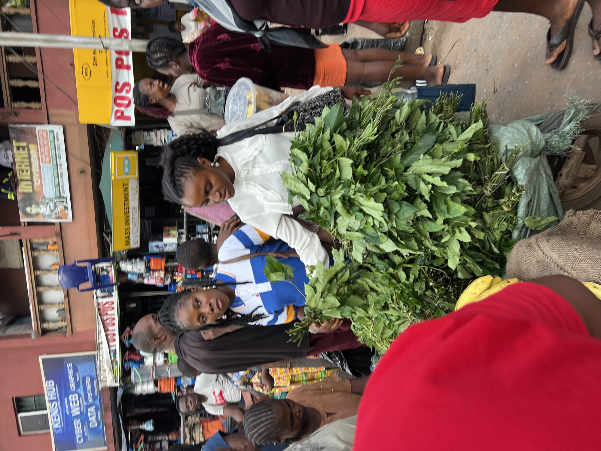 This is a medium shot photograph taken outdoors at a busy market in Nkpor, Nigeria. The foreground is dominated by a large pile of leafy green vegetables, likely produce for sale, bundled and resting on a sack. To the right of the vegetables, a person wearing a bright red skirt and a dark top is visible, with their legs and feet in sandals. To the left of the vegetables, a person in a red shirt is partially visible from behind.
In the mid-ground, several people are interacting. A woman with braided hair is holding some of the vegetables, looking towards the left of the frame. She is wearing a blue and white striped polo shirt. Another woman, with darker braided hair, is to her left, facing towards the right and appears to be speaking. She is wearing a light-colored, ruffled blouse. Behind them, a man in a dark t-shirt is visible. Further back, more people and market stalls are present.
The background reveals more of the market environment with various signs and structures. A prominent blue sign reads "KENIS HUB" with text below it about "CYBER WEB GRAPHICS" and "DATA". Another sign above the market stalls has "INTERNET CYBER" printed on it. Yellow signs with red and white text are also visible. The scene is crowded, indicative of a typical African market setting. The overall lighting suggests it is daytime.