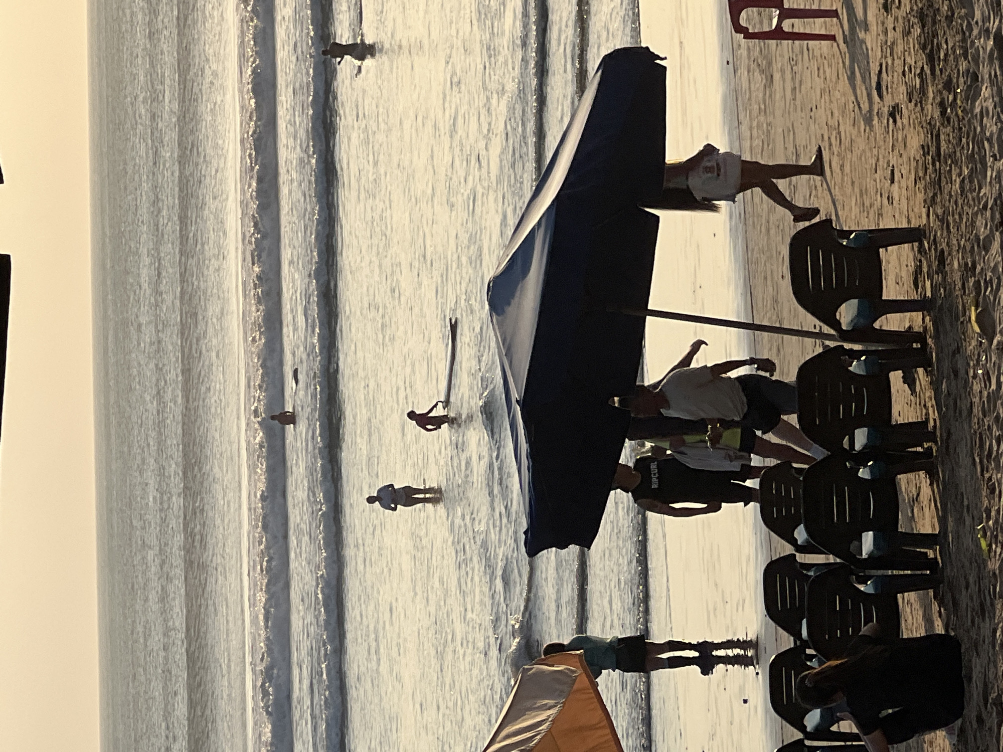 In Kuta, Indonesia, on a sandy beach with gentle waves, several individuals are engaged in various activities. In the foreground, a dark blue beach umbrella partially obscures a group of people. A person in a black t-shirt with "RIPCURL" visible is standing. Nearby, a woman in a white top and a dark skirt walks away from the camera. A few empty plastic chairs are set up on the sand.
In the middle ground, several people are in the shallow water. One person is standing, another is kneeling beside a surfboard, and a third person is further out in the water. Two more individuals are visible further out, one on a surfboard.
In the background, the ocean extends to the horizon under a hazy, light-colored sky, suggesting either sunrise or sunset. The overall scene depicts a relaxed beach atmosphere with people enjoying the water and the shore.