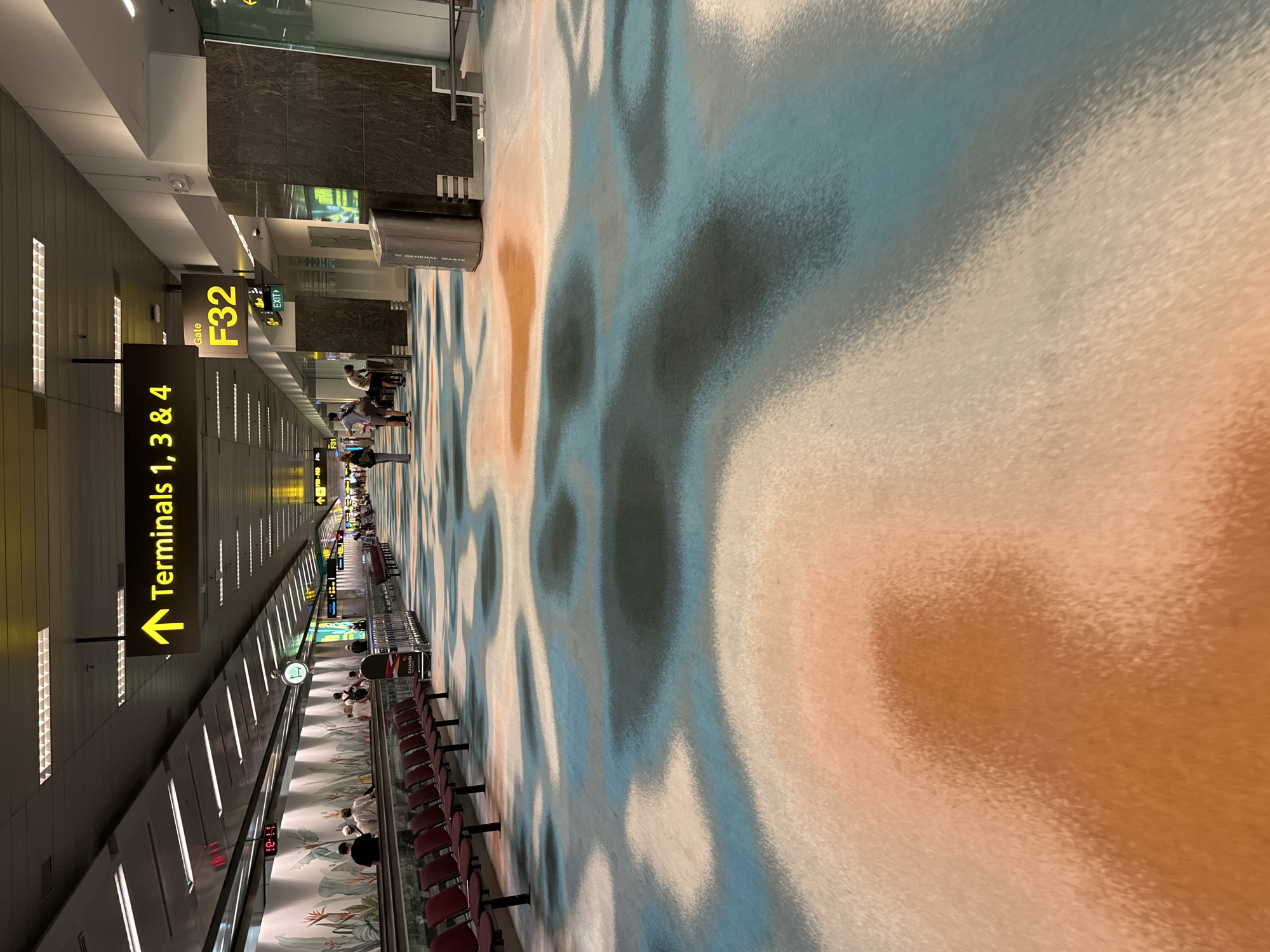 The image depicts a wide airport terminal concourse, likely in Singapore Changi Airport, given the visible "CHANGI" branding on a luggage trolley. The floor features a large, abstract, water-like pattern with swirling colors of orange, white, and various shades of blue and gray. Seating benches, colored a deep burgundy, are arranged along the left side of the concourse. People are seen walking and sitting, with some riding on a travelator in the background. Overhead, directional signage indicates "Terminals 1, 3 & 4" and "Gate F32," further confirming an airport setting. The environment is well-lit by artificial lighting from the ceiling.