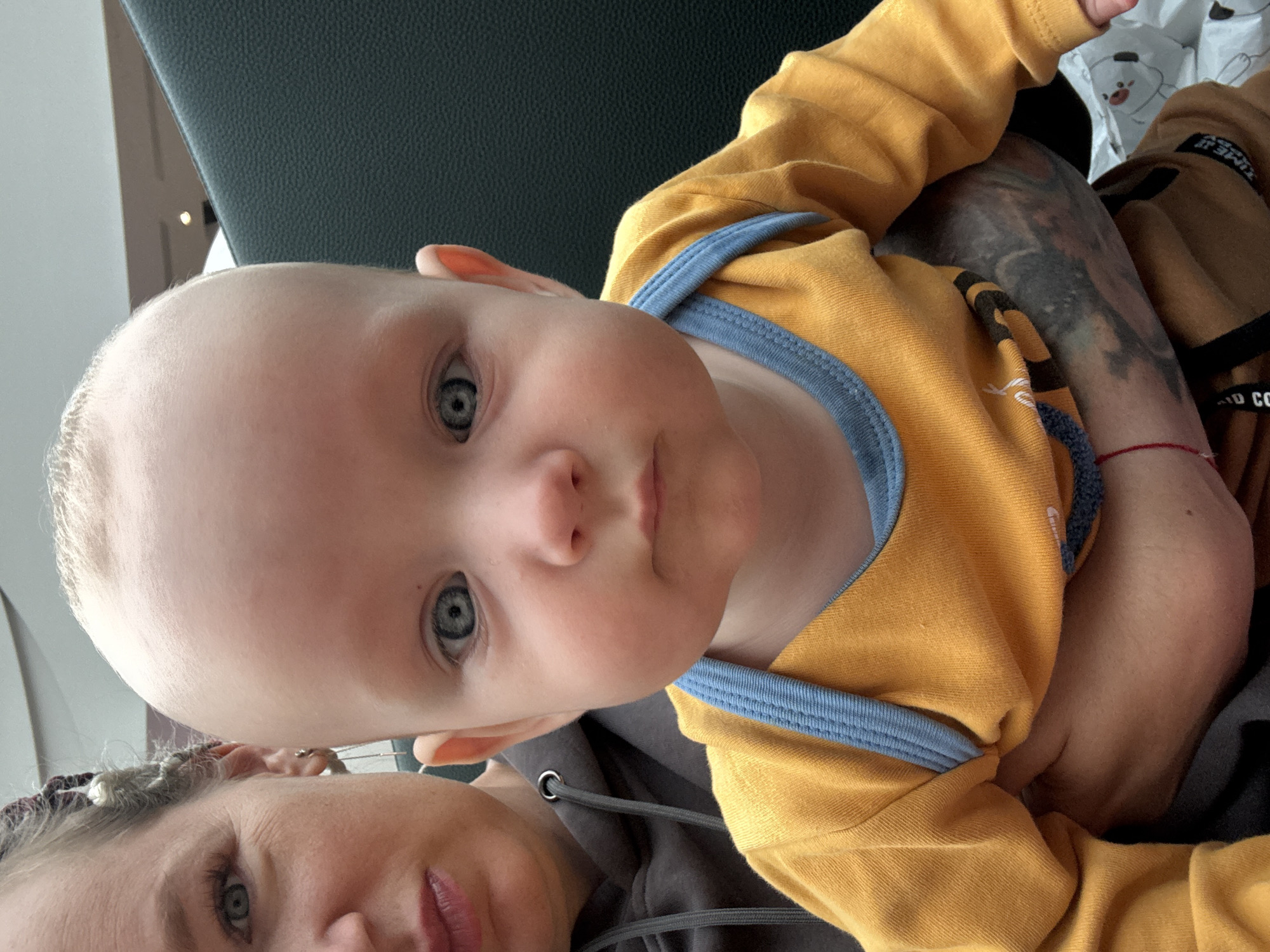 An adult and a baby are positioned in the foreground, with the baby's face taking up most of the frame. The baby has fair skin, light hair, and blue eyes, and is wearing a yellow long-sleeved shirt with a blue trim. The adult, to the left of the baby, has blonde hair styled in braids and is wearing a gray hooded sweatshirt. The background shows a dark, textured surface, possibly upholstery, and a glimpse of a ceiling with lighting. There is no indication of the location being Moscow, Russia, or any specific city. The scene appears to be an indoor setting, possibly a public space like a waiting area or transport.