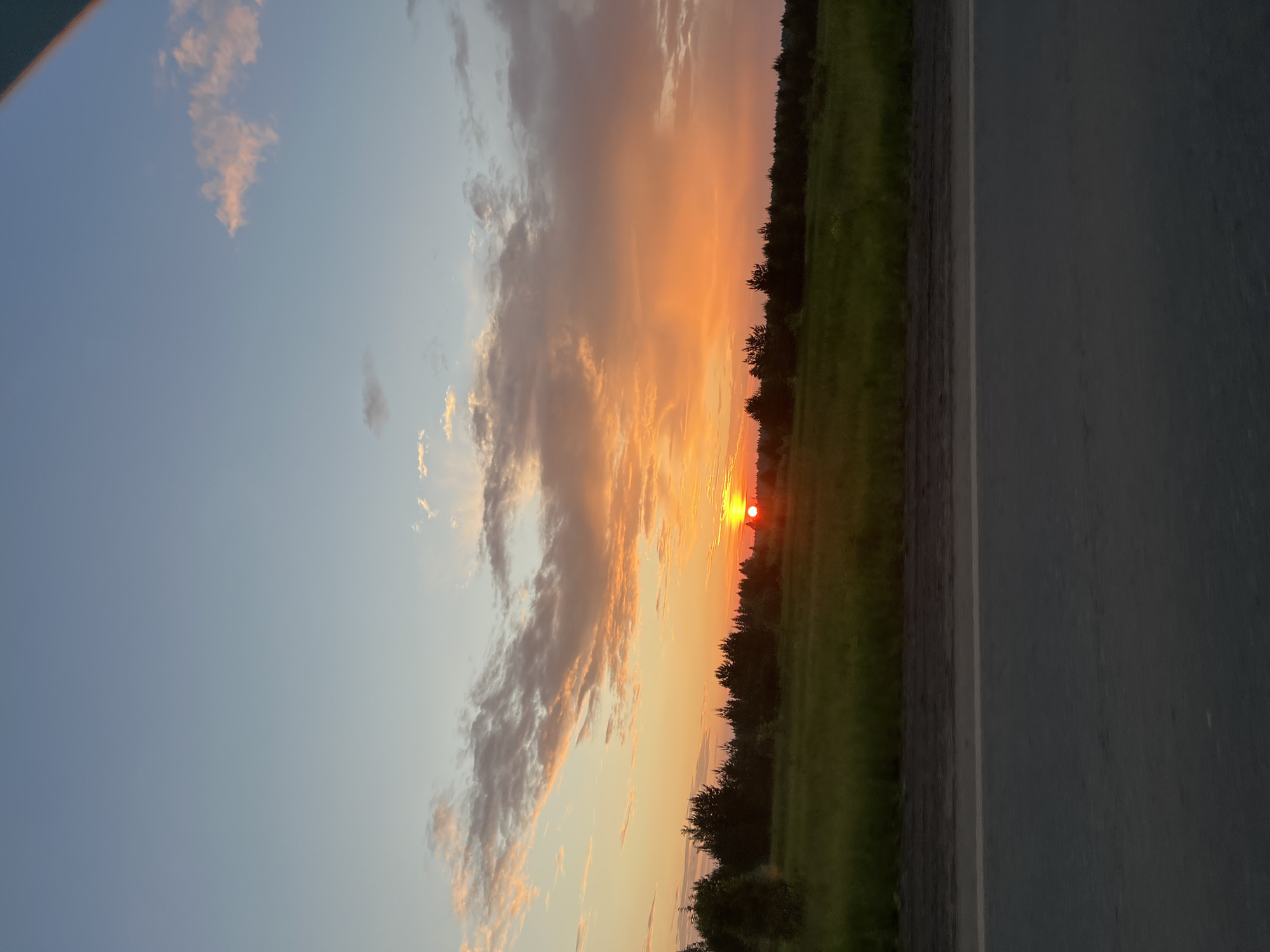 This image depicts a sunset scene in the vicinity of Поварёнки, Russia. The sky is filled with a gradient of colors, transitioning from a pale blue at the top to vibrant oranges and yellows near the horizon, illuminated by the setting sun. Wispy clouds, tinged with pink and orange, are scattered across the sky.
Below the sky, a line of dark green trees forms a dense treeline across the middle ground, suggesting a forest or woodland. In front of the treeline, a bright green field stretches out, appearing lush and slightly illuminated by the sunset.
In the foreground, a paved road runs horizontally across the bottom of the image. The road is dark gray, with a lighter gray stripe indicating a lane marker. The perspective suggests the photo was taken from a moving vehicle on this road, looking towards the sunset. No people or specific structures are visible in the scene.