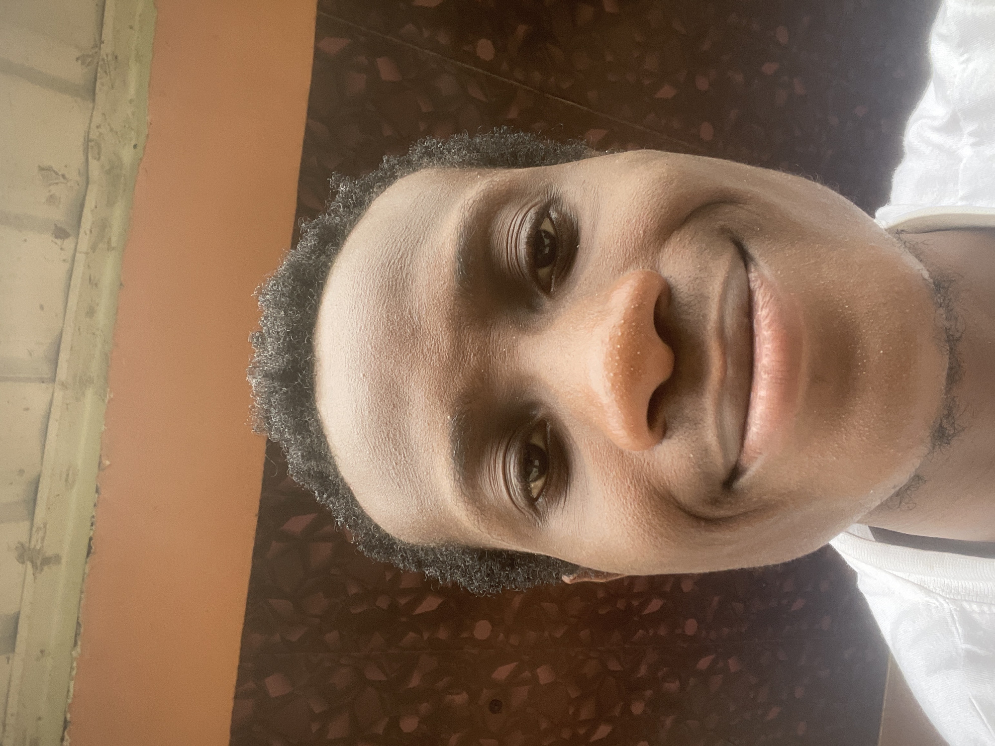 The image shows a close-up, low-angle selfie of a young Black man. He is wearing a white shirt. His head is tilted slightly back, and he is smiling. The background is a textured, dark brown wall with some lighter colored patterns, and above that, a band of orange and a lighter, weathered ceiling or overhang. The image appears to have been taken outdoors, possibly in the city of Ihiagwa, Nigeria.