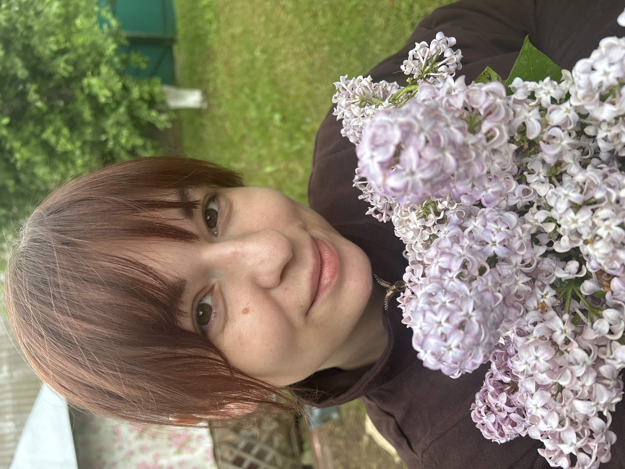 A woman with short brown hair and brown eyes is holding a bouquet of light purple and white lilacs. She is wearing a brown shirt or jacket. The background shows a green lawn and some trees. It is daytime. The exact location in Фоки, Russia is not discernable from this image.
