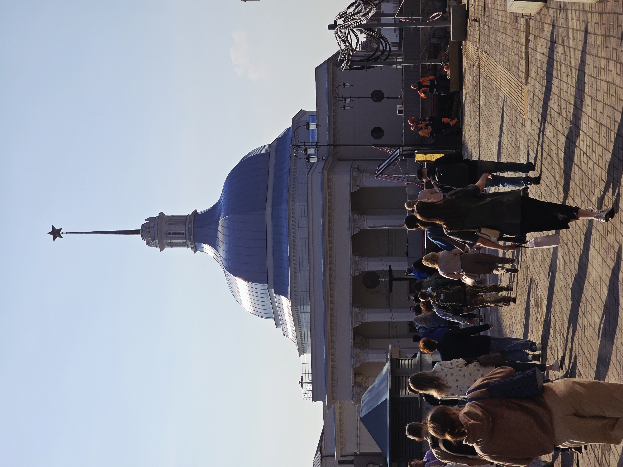 A low-angle, eye-level shot depicts a blue-domed building with a spire topped by a star against a clear, light blue sky, likely the Kazan Family Center, a civil registry building located in Kazan, Russia. A crowd of people stands on a paved brick walkway in front of the building, casting long shadows on the ground. Construction workers wearing orange vests and hardhats are visible near the building. Wires and scaffolding are present near the building.