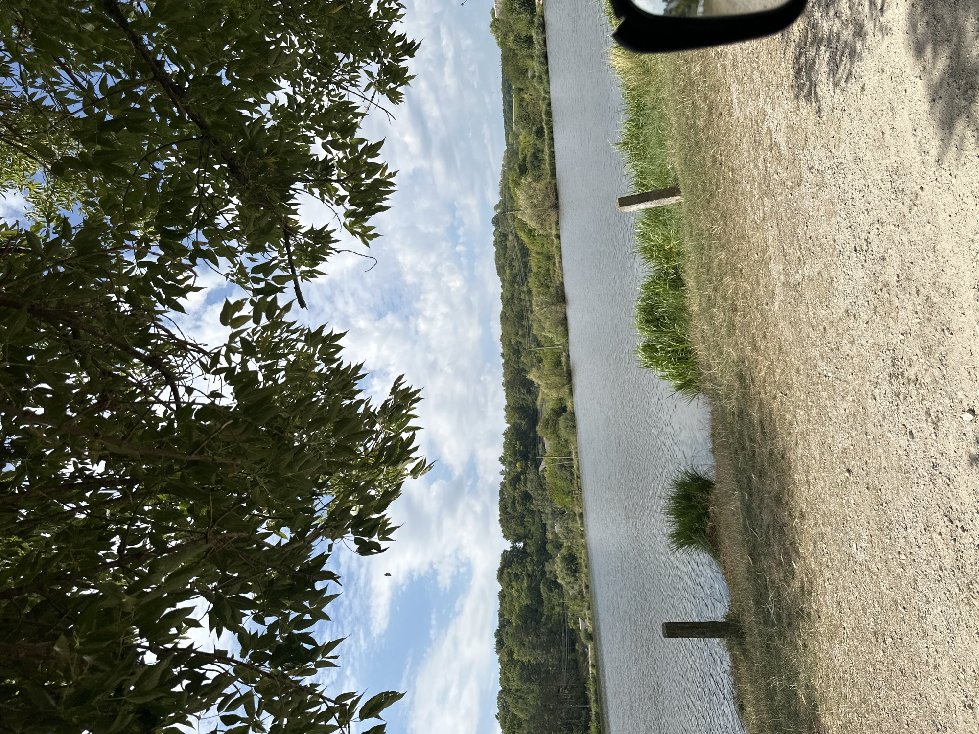 An outdoor, eye-level, full shot depicts a body of water with a tree in the foreground. The water is calm with ripples on the surface. The foreground is a dirt path with dried grass and weeds on the left and right. Two wooden posts are in the foreground, one on the left and one on the right. Green grass and reeds are visible on the edge of the water. A dense line of trees and vegetation forms the background. The sky is blue with white clouds. This scene is in the city of Makiv, Ukraine.