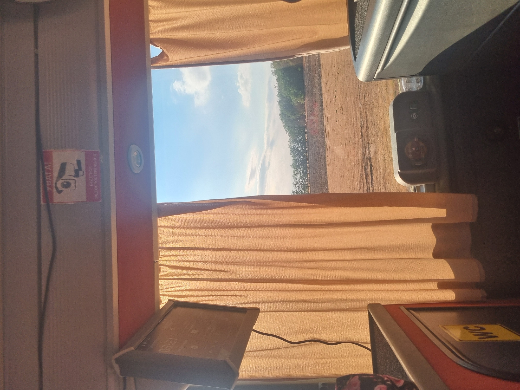 The image shows the interior of a bus, looking out a window. The window is partially covered by a tan curtain. Through the window, a dry, agricultural field is visible, with a line of trees in the distance under a cloudy blue sky. Inside the bus, there is a small screen on the left, a WC sign on a cabinet below it, and a control panel with several buttons and an outlet on the right. A sign with a camera icon and text in Ukrainian is visible at the top of the frame. The setting appears to be during daylight hours. The location is not specified in the image, but it is stated to be in Berettyóújfalu, Hungary.