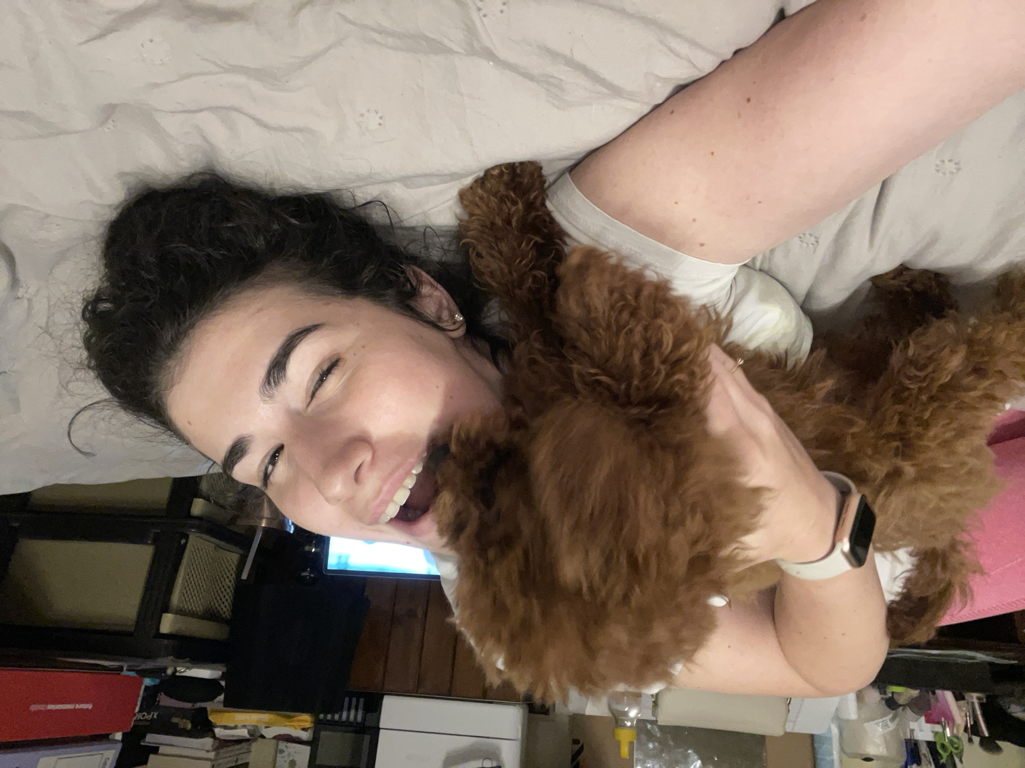 A young adult female is lying on a bed with a light gray patterned comforter. She has dark, curly hair pulled back in a messy bun, fair skin, and appears to be smiling with her eyes squinted slightly. She is wearing a light-colored t-shirt. A small, brown, fluffy dog is positioned on her chest and is being held by her arms. She is wearing a white smartwatch on her left wrist.
In the background, a cluttered area can be seen, including a dark-colored dresser or cabinet, a television screen with a blue light emanating from it, and various objects such as books, boxes, and miscellaneous items piled on the floor.
The location is not discernible to be Sofia, Bulgaria based on the image.