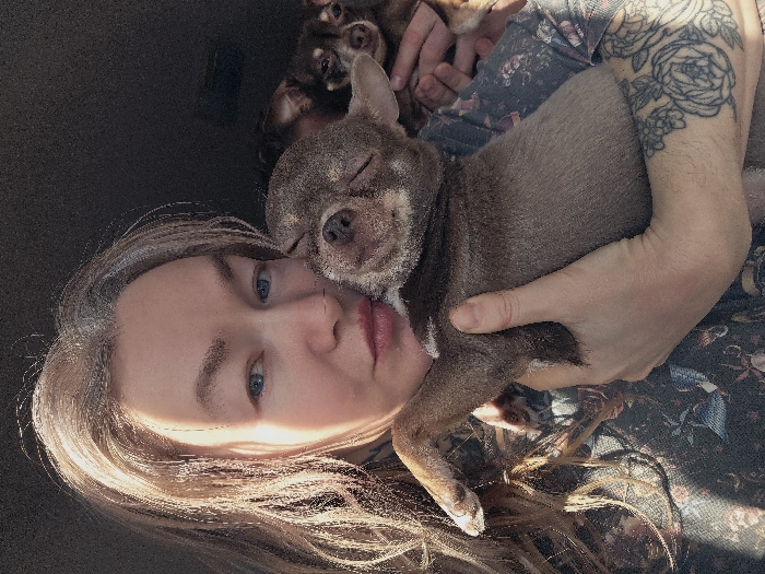 An individual with long, light brown and gray hair, blue eyes, and fair skin is depicted in a close-up, indoor photograph, holding two small dogs. The person wears a dark, patterned garment and has a floral-themed tattoo on their left forearm. They hold a small, short-haired, brown dog with lighter fur on its muzzle and paws, which is resting its closed-eyed head against the person's left cheek. A second, smaller brown dog is partially visible in the upper right background, looking towards the camera. The background is dark and out of focus, suggesting an interior space, possibly a vehicle. Natural light illuminates the left side of the person's face and hair. No specific identifying features are present to determine a precise location within Saint Petersburg, Russia.
