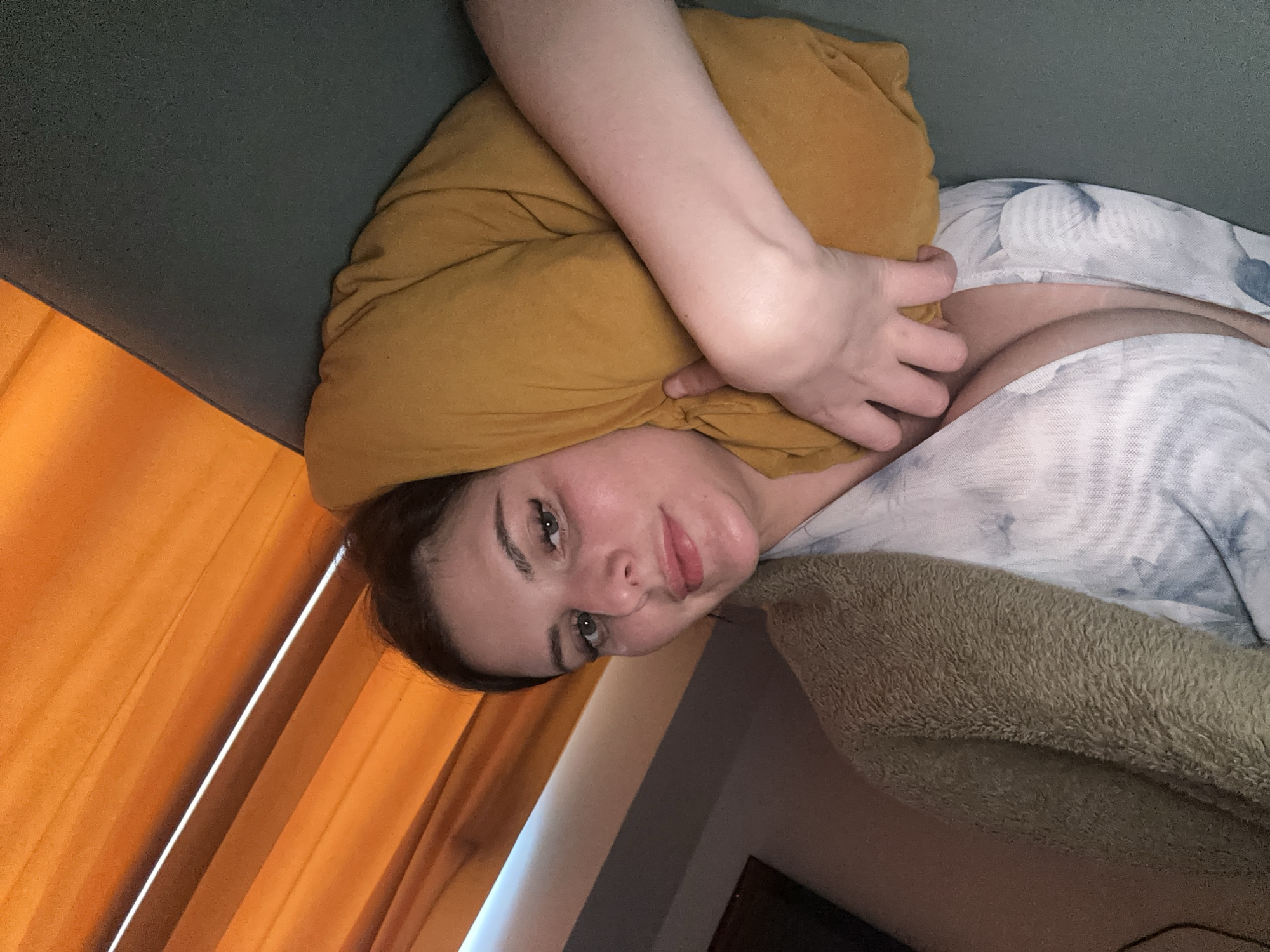 A young woman with light skin, dark hair, and green eyes is lying on a gray sofa, propped up by a yellow pillow. She is wearing a white and blue patterned robe. She has on dark eye makeup and her lips are slightly parted. Her left arm is resting on the pillow, and her right hand is tucked beneath it. Behind her, orange curtains are visible, and a fuzzy, light brown blanket is draped over her left shoulder. The scene appears to be indoors, possibly in a living room.