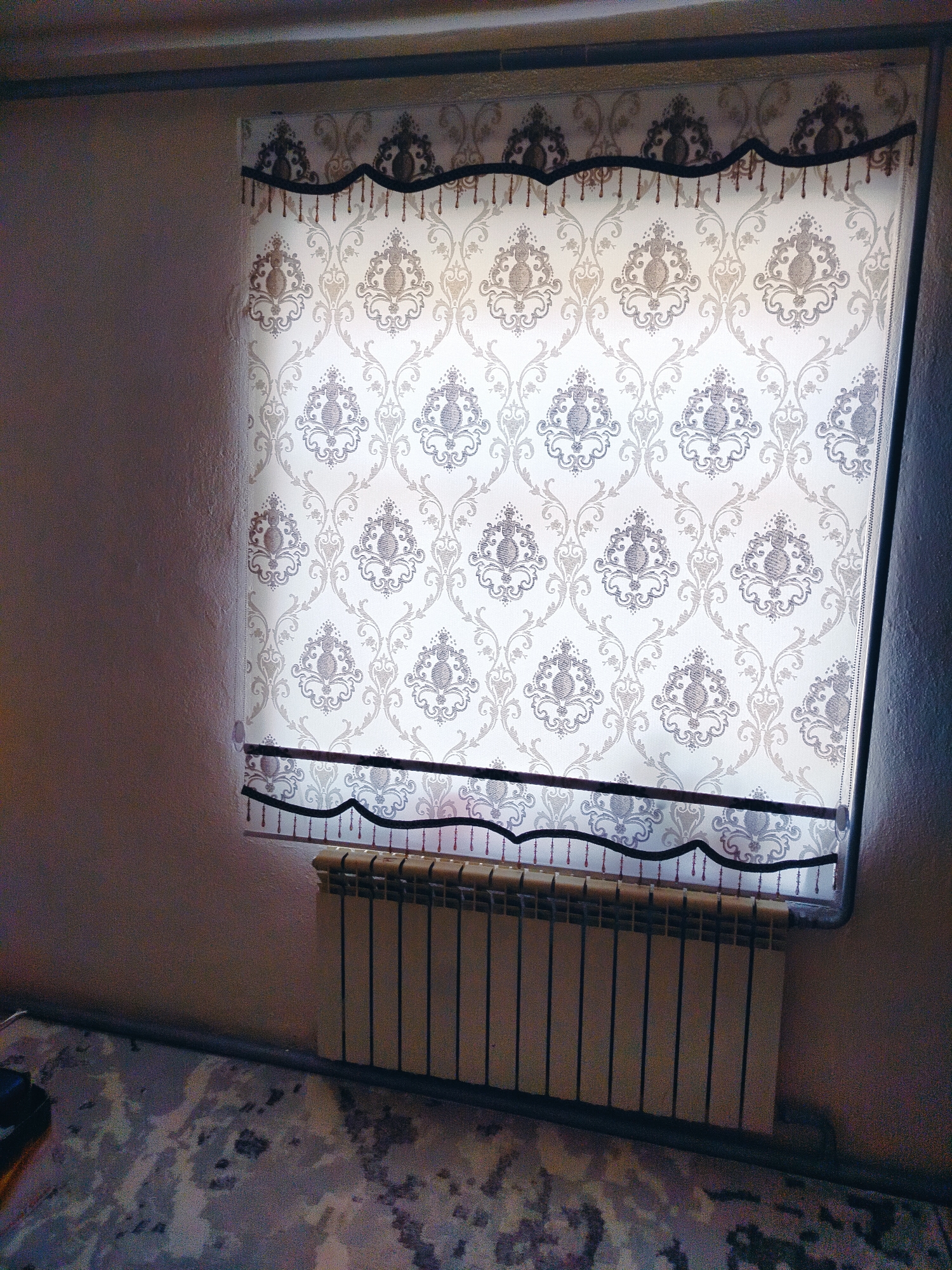 The image shows a window with a patterned roller blind in a room. The blind has a decorative trim at the top and bottom, featuring dark fabric with beading and tassels. Below the window is a radiator with vertical fins. The wall to the left of the window is textured and appears to be a neutral color, possibly painted or wallpapered. The floor has a patterned carpet with abstract designs in shades of gray, beige, and dark blue. The lighting in the room is dim, casting shadows on the wall. The overall scene is domestic, with no people or immediate signs of activity. The location is described as Көлкент, Kazakhstan, suggesting this is a residential interior in that region.