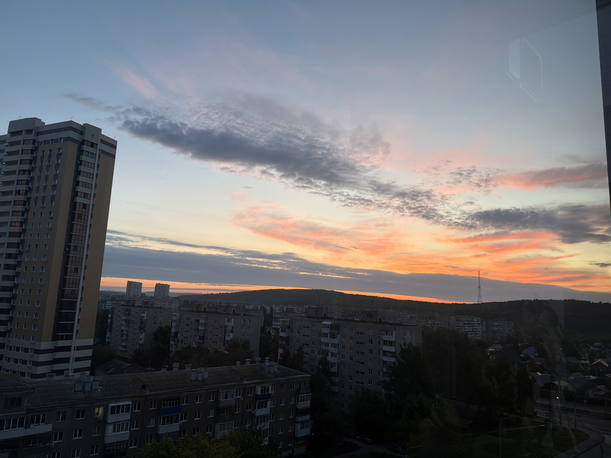 This is a high-angle, medium shot of a cityscape during sunset, taken from an interior vantage point, likely a window with reflections visible. The sky is filled with a gradient of colors, transitioning from a pale blue at the top to vibrant oranges and pinks near the horizon, with scattered clouds.
Dominating the left side of the frame is a tall, modern apartment building with a light beige and grey facade, featuring numerous windows. To the right and in the mid-ground, a dense collection of older, grey apartment blocks are visible, characteristic of Soviet-era architecture, with their distinctive balconies and window arrangements. Further in the distance, a low, forested hill forms the horizon, with two prominent, taller buildings silhouetted against the colorful sky. A telecommunications tower is also visible on the horizon.
The foreground shows the tiled roofs of some of the older buildings, and the bottom right includes a glimpse of a road with streetlights and some smaller houses. The reflections in the window are indistinct but suggest the interior of a room. There are no visible people or animals. The overall impression is a vast urban landscape bathed in the warm, atmospheric light of a setting sun.