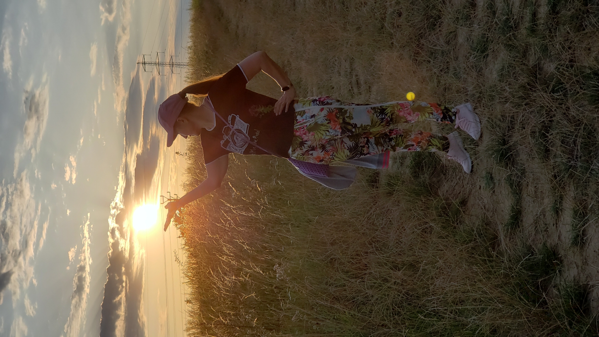 A woman stands in a field of dry grass at sunset, holding the sun in her open palm. The sun is setting behind a band of clouds, casting golden light across the scene. The woman is wearing a dark t-shirt with a camera graphic, colorful floral patterned pants, and a purple baseball cap. She has a bag slung across her shoulder. In the background, a power line pylon is visible against the sky. The location is the vicinity of Prešov, Slovakia.