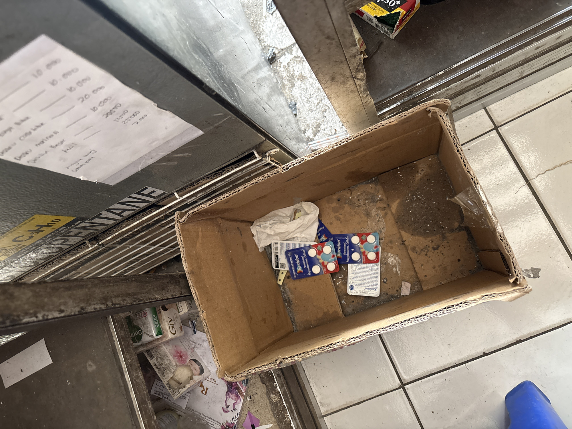 A cardboard box containing two blister packs of Saridon tablets, a crumpled white cloth, and what appears to be a small paper is placed on a dirty floor. In the background, a metal cabinet is visible with some items on its shelves, including a box of Dettol soap and other products. A yellow sign with "C5H10" is also visible on the cabinet. The floor is tiled with white squares and grout lines, and some debris is scattered around. The scene suggests a storage or discarded area, possibly at a shop or a warehouse in Palimanan, Indonesia.