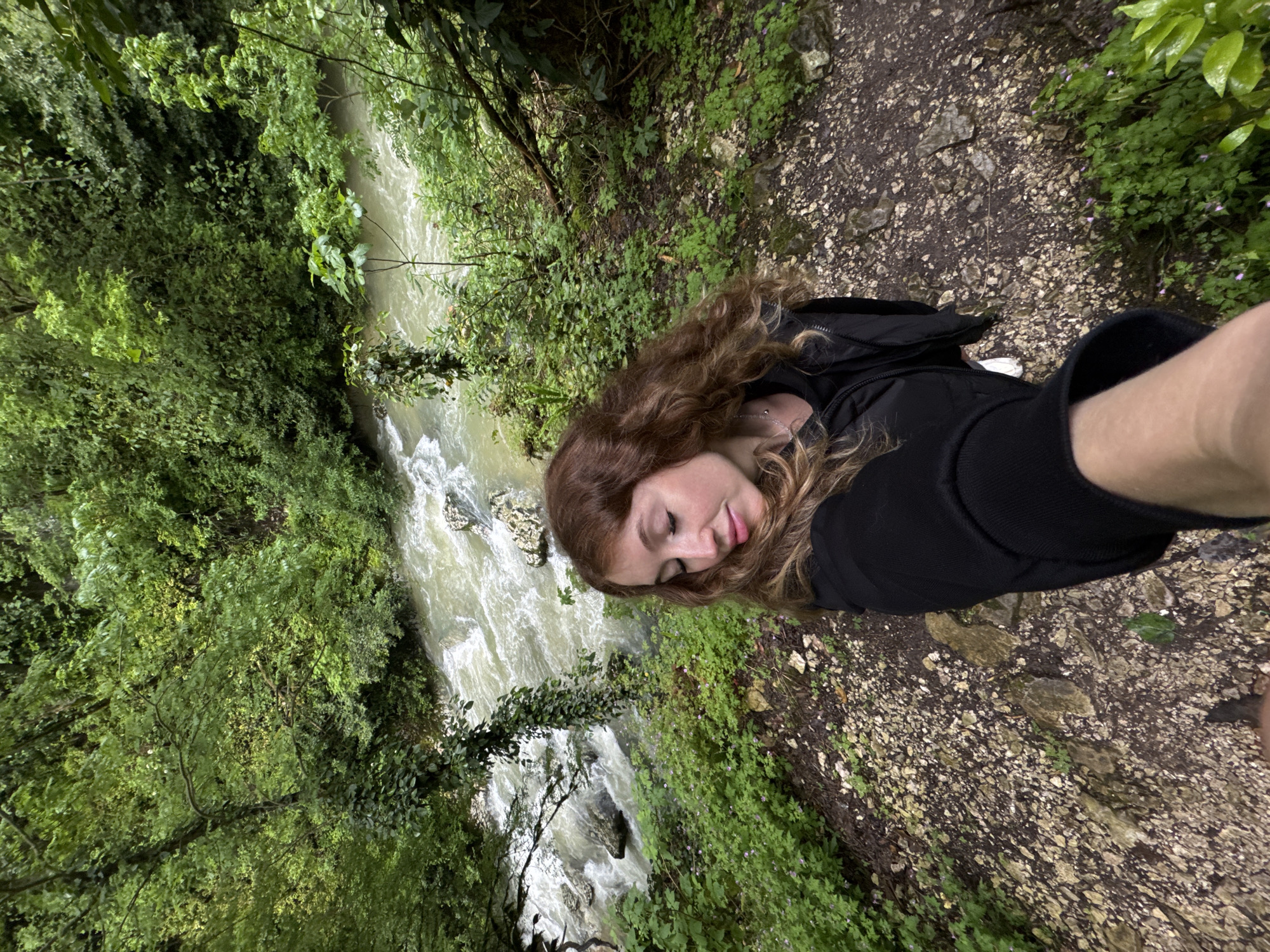 A person is standing on a dirt path beside a fast-flowing river in a forest. The river is on the left side of the image and the forest extends upwards, filling the left of the frame. The path is covered in loose rocks and vegetation, and takes up the right portion of the image. The person is wearing a black jacket and appears to be taking a selfie. The location may be in Tsabal, Abkhazia, based on information provided.