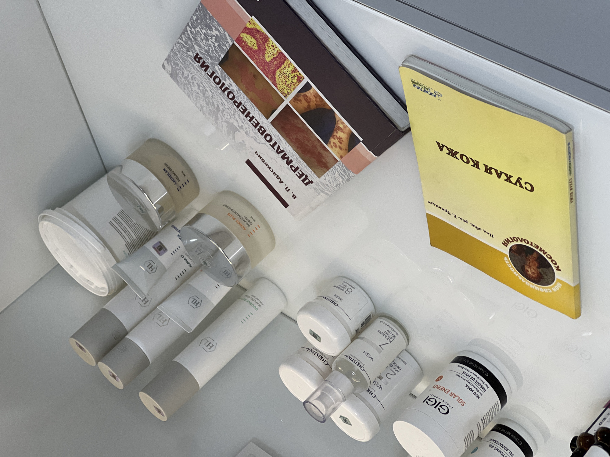 The image shows a top-down view of a shelf with various skincare and cosmetic products, along with two books. The products include items from brands such as "HL," "Christina," and "Gigi Laboratories." Some of the products are labeled as "Peeling Cream," "Daydream," "Eye & Neck Lifting Serum," and "Mud Mask." The books are titled "Dermatovenerology" and "Dry Skin: Cosmetology." The scene is a well-lit, modern environment, likely a cosmetics store or a medical spa. There is no specific information available in the image to definitively indicate the location is in Yerevan, Armenia.