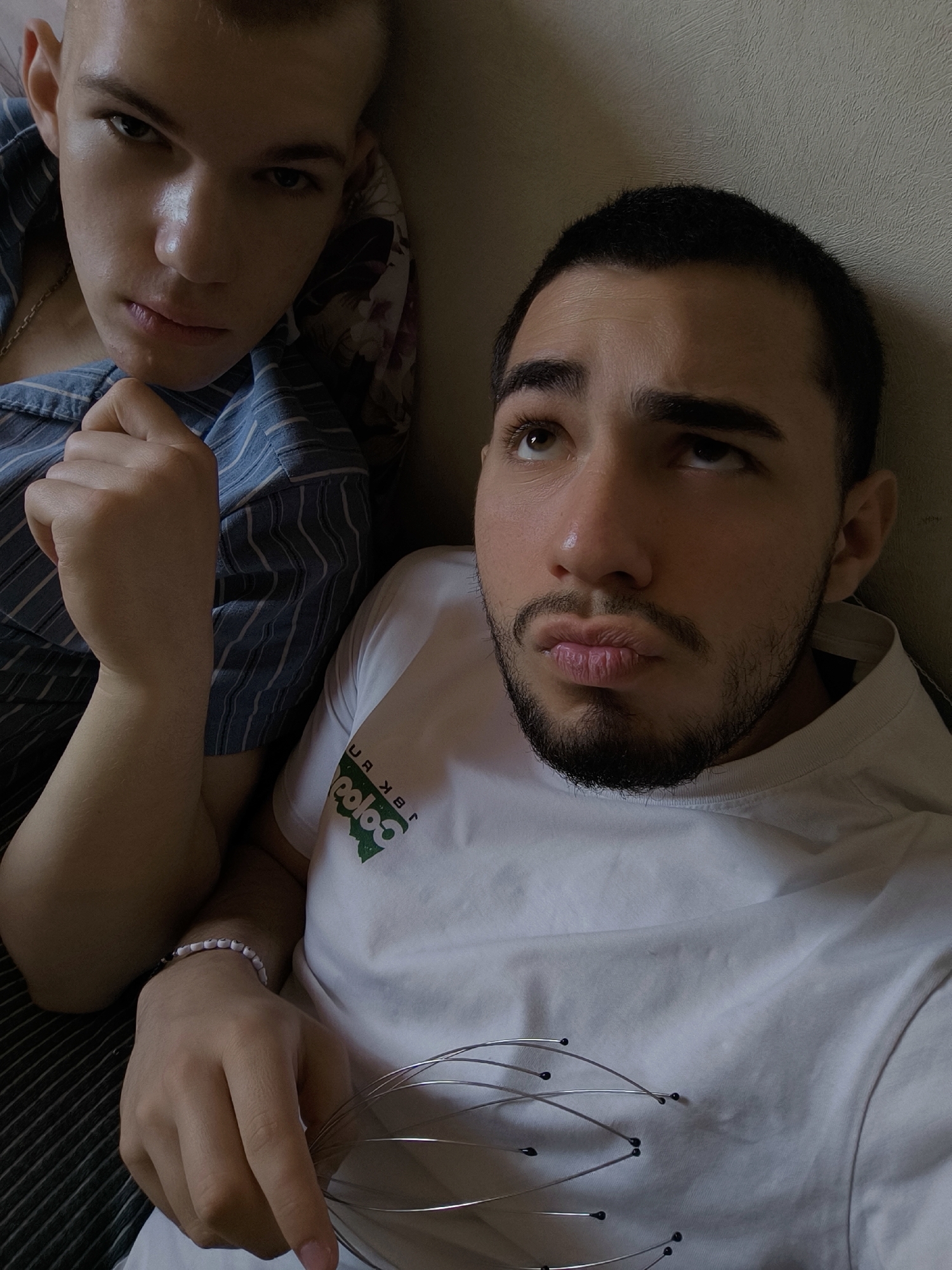 Two young men are pictured in a close-up, casual indoor setting, likely in Schiltigheim, France. The man on the left has short, light-colored hair and is wearing a blue and white striped shirt with a faint gold chain visible. He looks directly into the camera with an intense, serious expression, his right hand clenched into a fist. The man on the right has short dark hair, a beard, and a mustache. He is wearing a white t-shirt with a green, partially reversed logo that reads "coloc" (appearing as "ooloc" due to the angle) and what appears to be reversed Cyrillic text above it. He looks upwards with a pouting expression, holding a silver wire head massager with black tips in his left hand, which also features a white beaded bracelet. The background consists of a light-colored wall or surface and a floral patterned fabric, possibly a pillow or blanket. The lighting is soft and diffused, creating a relaxed yet expressive atmosphere.