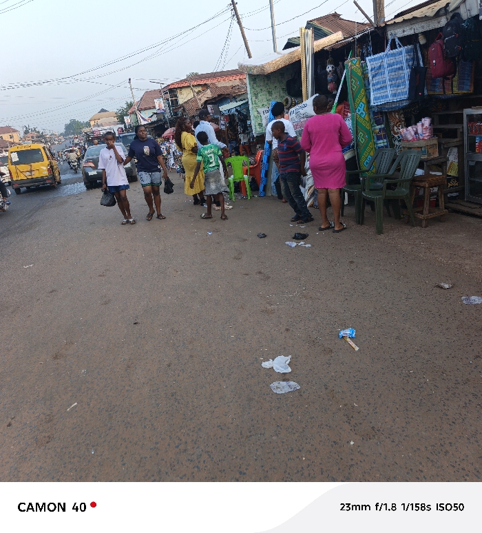 A bustling street scene is captured with several people traversing the road. A taxi, painted in yellow, is visible on the left. People stand near shops on the right, some looking at merchandise. The street itself shows signs of litter. The perspective offers a broad view of the surroundings, capturing both the human activity and the urban environment.