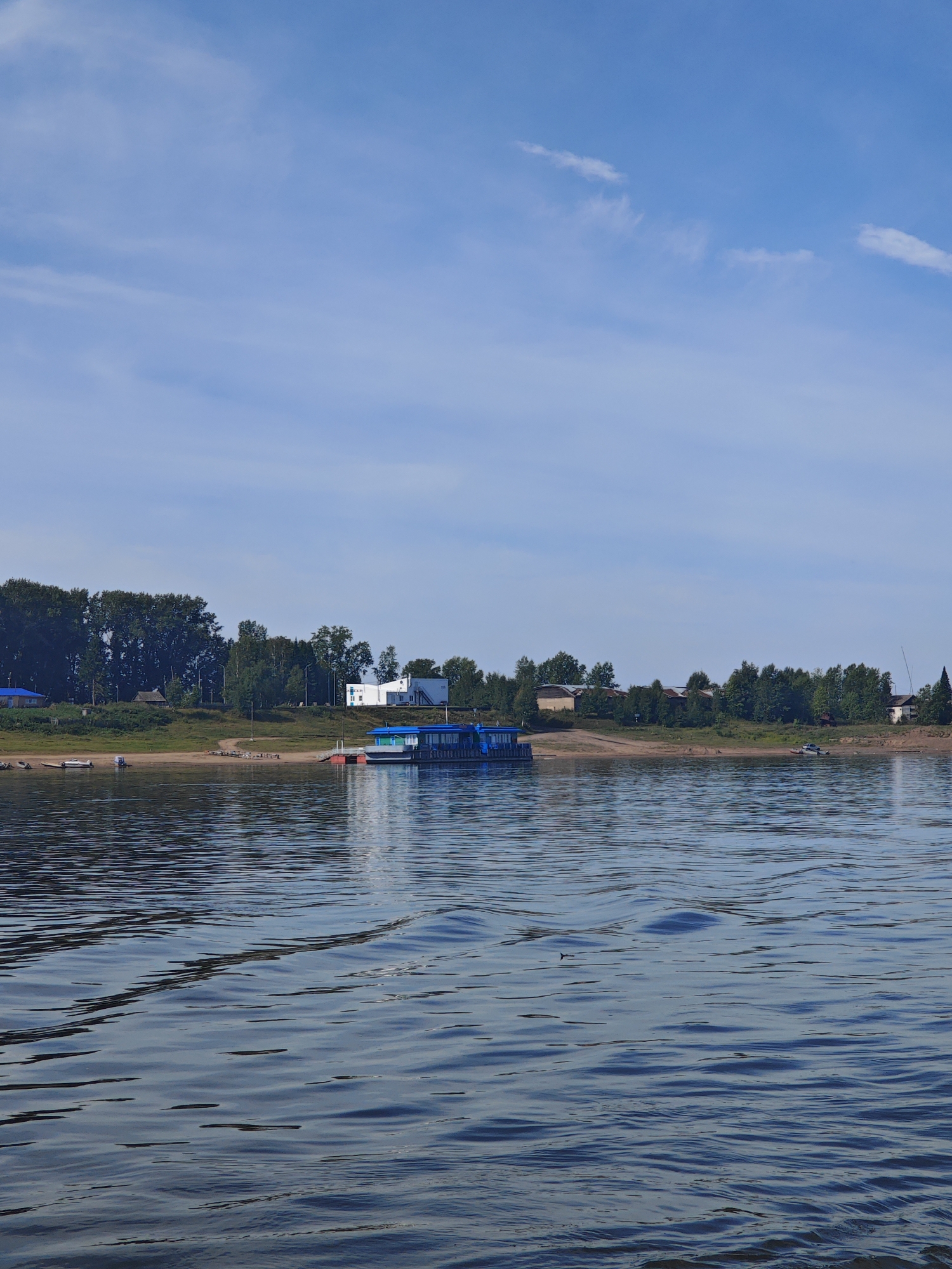 The image shows a wide river with gentle waves under a partly cloudy blue sky. On the far bank, a line of trees forms a dense forest. Along the edge of the river, there is a sandy embankment with a few scattered buildings and a larger, modern white building. A blue and white boat is docked at a pier. In the foreground, the water is dark and reflective, with sunlight creating ripples and patterns on its surface. The scene appears to be a tranquil, daytime view of a riverbank settlement. There are no people visible in the image. Based on the limited visual information, it is not possible to definitively determine the specific location within the city of Yartsevo, Russia.