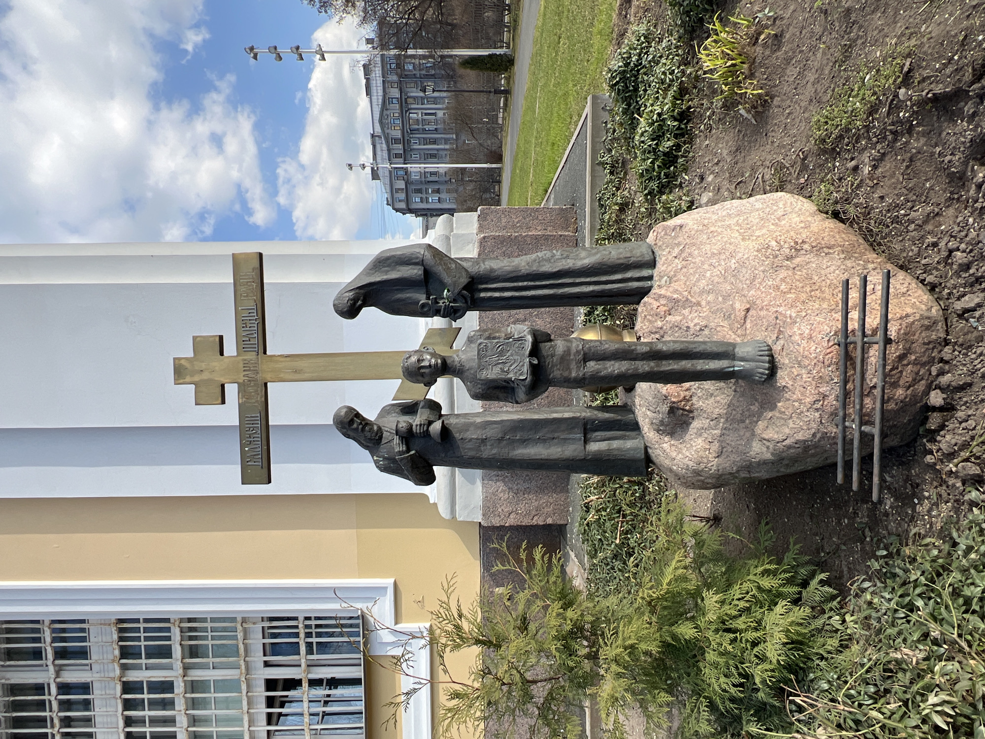 The image captures the Monument to the Victims of Political Repression (Solovetsky Monument) in Troitskaya Square on Petrogradsky Island, Saint Petersburg, Russia.
The central subject is a bronze sculptural group situated on a large, irregularly shaped pinkish-brown granite boulder. Three stylized figures are depicted: a barefoot male youth stands centrally, holding an open book with an emblem; to his left, an older bearded male figure in a long robe holds a chalice-like object; to his right, a cloaked female figure, with her head bowed, holds a small crucifix or religious artifact. Behind these figures, a brass-colored Orthodox cross is erected, bearing the Cyrillic inscription: "БЛАЖЕННЫЕ ИЗГНАННЫЕ ПРАВДЫ РАДИ" (Blessed are those who are persecuted for righteousness' sake). At the base of the boulder, in front of the youth, three horizontal dark metal bars are visible.
The monument is set within a garden bed of dark soil, interspersed with sparse green ground cover and a small, vibrant green coniferous shrub positioned to the left.
In the immediate background to the left, a multi-story building with a light yellow facade and numerous windows, each secured by white metal grilles, is visible, consistent with the House of Political Prisoners. To the right, beyond a wide expanse of green parkland with bare trees and a paved pathway, a large, grey multi-story building featuring classical architectural elements such as columns and cornices stands in the distance. Several tall utility poles with light fixtures and a traditional black street lamp are visible within the park. The scene is illuminated under a partly cloudy blue sky.