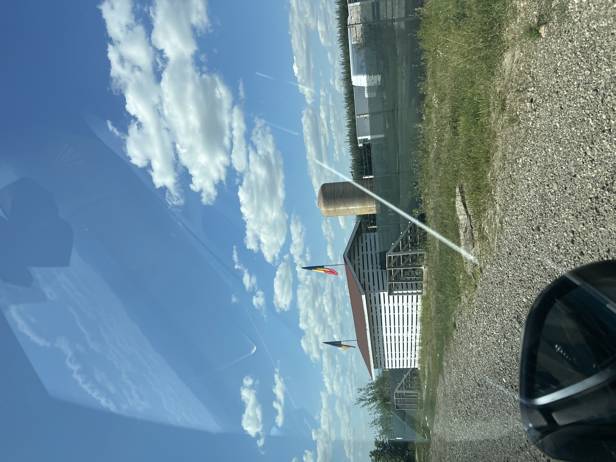 The image depicts an outdoor scene on a clear, sunny day with a bright blue sky filled with scattered white clouds. In the foreground, a gravel path leads towards a white, slatted structure resembling a shed or small building, topped with a brown roof. Two flags, one with red, yellow, and black stripes and the other with black, yellow, and red stripes, are flying from poles on either side of the building. To the right of the building, a large, cylindrical, tan-colored tank stands. Beyond this, a green mesh fence encloses an area with some white rectangular objects, possibly storage containers or building materials, and a line of trees in the distance. The environment appears rural or semi-rural, with patches of green grass and some smaller white wildflowers visible. Reflections on what appears to be a window or car windshield are present in the upper portion of the image. The scene is devoid of people and indicates a static environment.