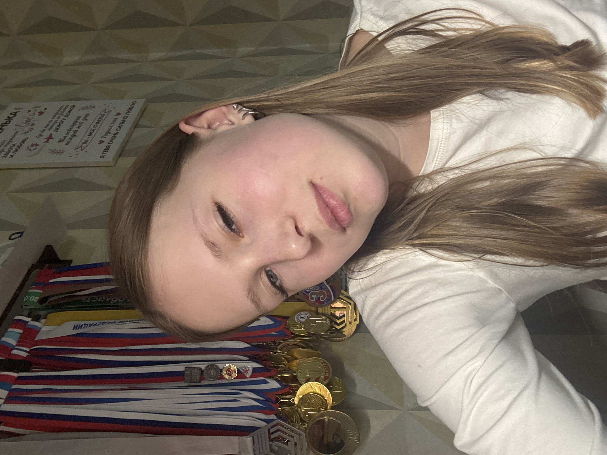 A young individual, likely female, with long, light brown hair, is pictured lying down, looking towards the camera with a relaxed expression. She is wearing a white long-sleeved top. The background features a geometric patterned wall and a decorative plaque.
Prominently displayed in the foreground and beside the person is a large collection of medals. Many of these medals are adorned with ribbons in the colors of the Russian flag (red, white, and blue), indicating a connection to Russia. Some visible inscriptions on the medals include "ФЕДЕРАЦИИ" (Federation), "область" (region), "МЕСТО" (place), and specific placements like "1" and "3 МЕСТО" (3rd place), suggesting achievements in competitions or sports. There are also numerous gold-colored medals with various designs.
On the wall behind the person's head, a white decorative plaque features handwritten text in Russian. The messages express sentiments of love and affection, including phrases such as "ЗАБОТА И ЛЮБОВЬ" (Care and Love), "ТЫ - МОЁ СЧАСТЬЕ" (YOU - MY HAPPINESS), "Родная моя" (My dear/beloved), and "Я ТЕБЯ ОЧЕНЬ СИЛЬНО ЛЮБЛЮ" (I LOVE YOU VERY MUCH). Other visible text fragments include "ДОРОГУ домой" (The road home) and "Ты освещаешь каждый мой день" (You illuminate every one of my days).
The scene is indoors, likely in a personal setting, given the relaxed pose and the personal messages on the plaque. The presence of numerous medals suggests the individual has achieved success in various endeavors, possibly sports, aligning with the implied location of Chaikovsky, Russia due to the Russian language and flag colors.
