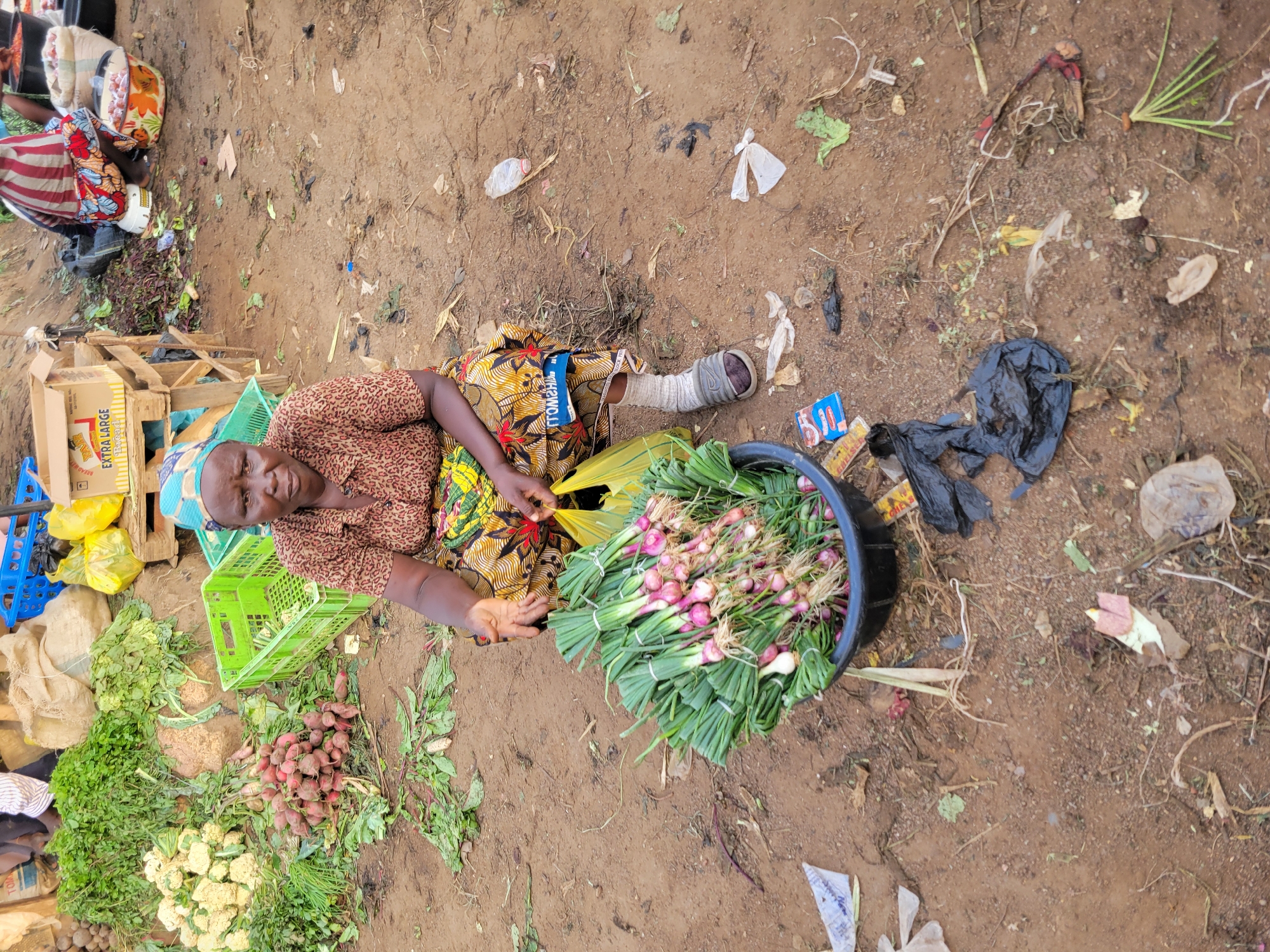 A woman sits on the ground in what appears to be a marketplace in Gura Top, Nigeria. She is wearing a patterned headscarf, a brown and patterned short-sleeved shirt, and a colorful wrap skirt. In front of her is a large black bowl filled with bunches of green onions. To her left, several green plastic crates are stacked, one containing white cauliflower. Behind her, a wooden stand holds boxes and sacks of produce, including radishes and leafy green vegetables. The ground is a mix of dirt and scattered debris, including plastic bags and food waste. The woman is looking towards the camera with a neutral expression.