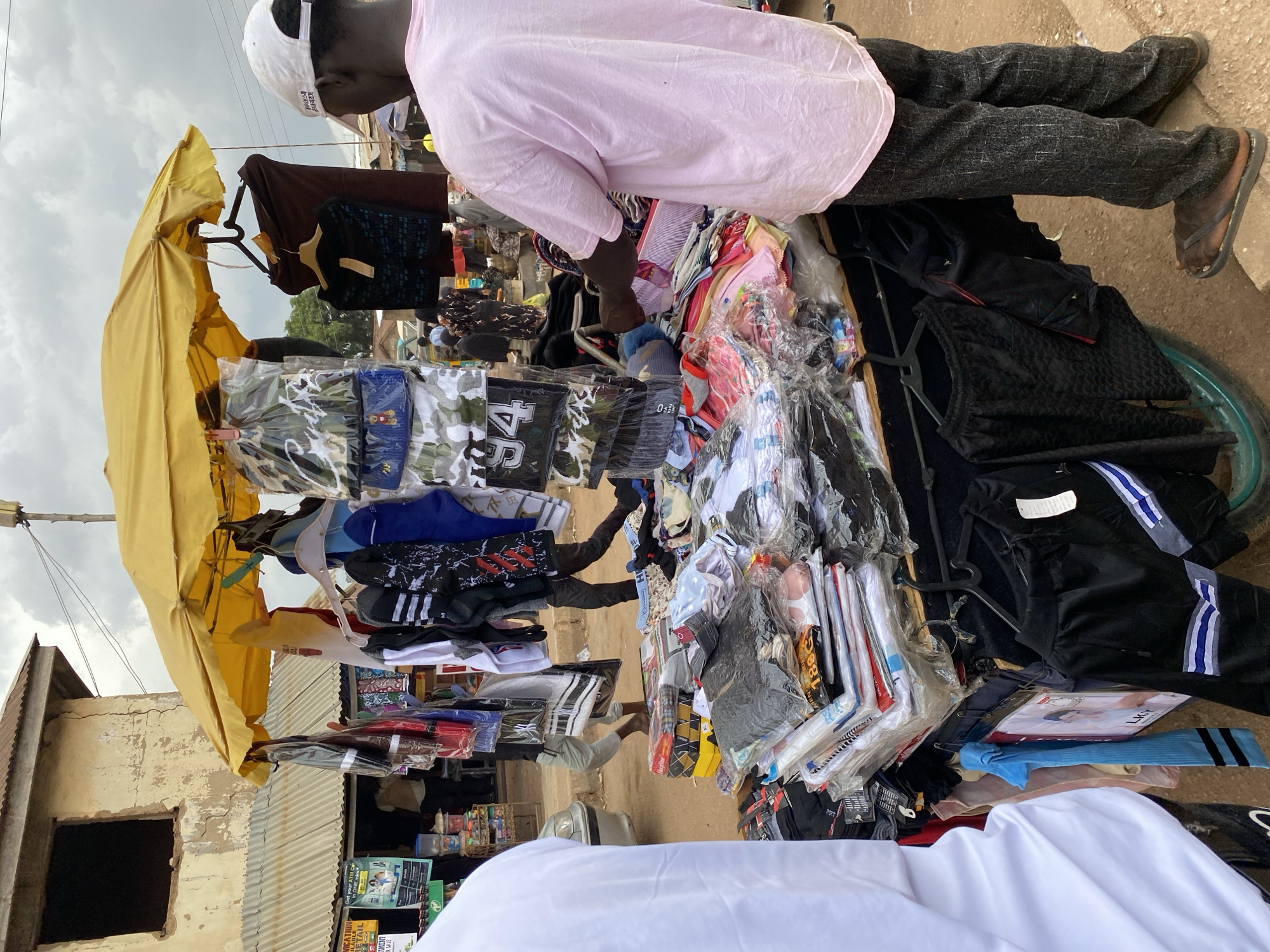The image depicts a street scene in Bukuru, Nigeria. A vendor, wearing a pink shirt, dark pants, and flip-flops, stands behind a mobile stand laden with clothing items. The stand is covered by a yellow umbrella. Assorted garments, including shirts, pants, and socks, are displayed on hangers and stacked on the stand. The vendor is examining or organizing the merchandise. In the foreground, a person wearing a white shirt is partially visible. The background includes buildings with corrugated metal roofs and posters.