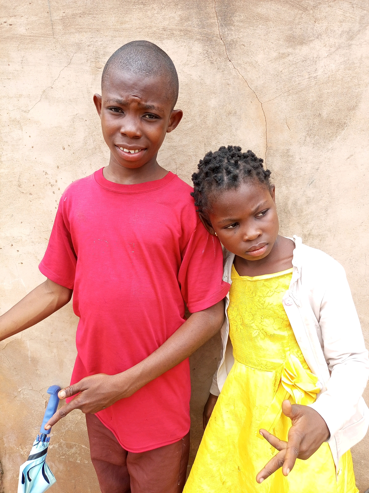 Two children, a boy and a girl, stand in front of a light brown, textured wall in Tempure, Nigeria. The boy, on the left, wears a red t-shirt and brown trousers. He has a close-cropped haircut and is looking at the camera with a slight frown. He holds a blue and white umbrella in his left hand. The girl, on the right, wears a yellow dress with a lace overlay and a light-colored cardigan. She has braided hair and is looking away from the camera with a thoughtful expression. Her right hand is raised, with two fingers extended in a gesture. The wall behind them appears to be made of stucco or concrete and has some visible cracks. The lighting suggests it is daytime.