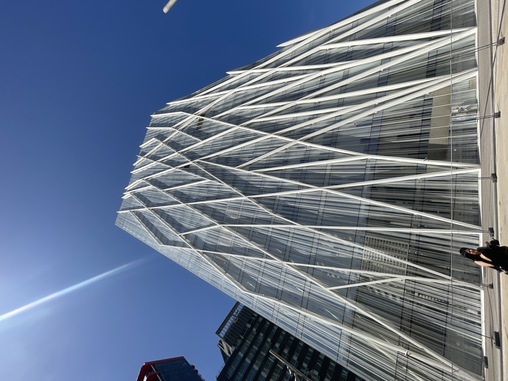 The image shows a low-angle view of a modern, glass-clad building in Barcelona, Spain. The building features a striking white exoskeleton design, creating a crisscrossing pattern on its facade. A clear, blue sky fills the background. A lens flare emanates from the lower-left corner. In the foreground, a person stands on the sidewalk, their face obscured by shadow. Adjacent buildings are visible at the base of the primary structure.