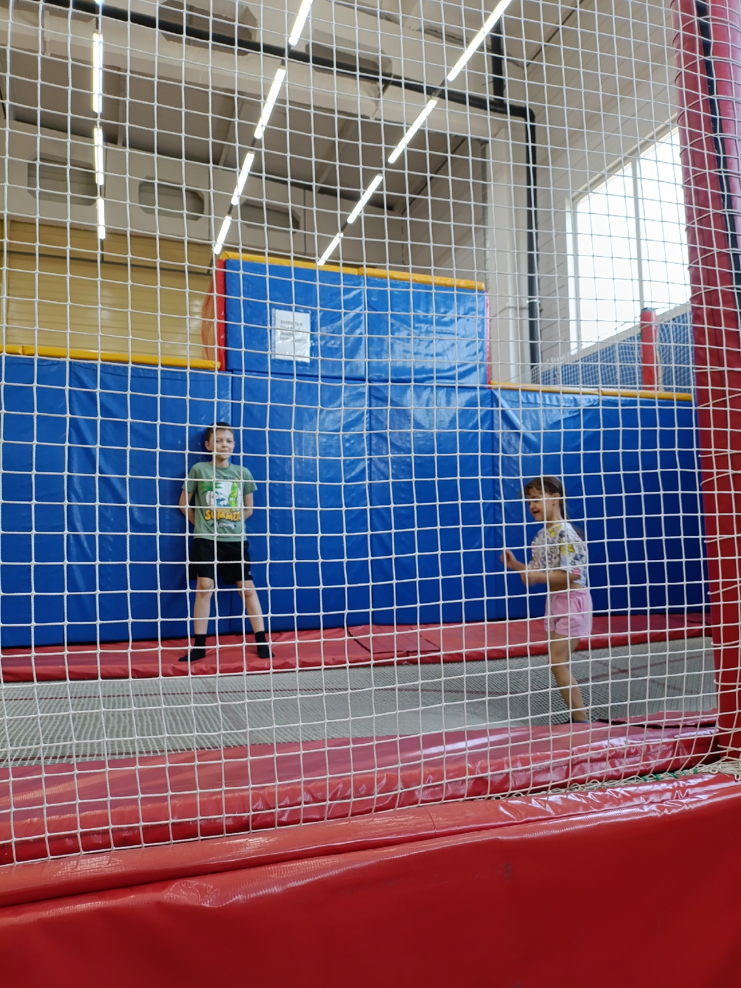 The image shows two children inside an enclosed trampoline park in what appears to be an indoor recreational facility. The location is Chaikovsky, Russia. A boy, wearing a green t-shirt and black shorts, is standing against a blue padded wall with red and yellow accents. A girl, wearing a patterned t-shirt and pink shorts, is to the right of him. Both are behind a white mesh safety netting with a red base. The trampoline park is brightly lit with overhead fluorescent lights.