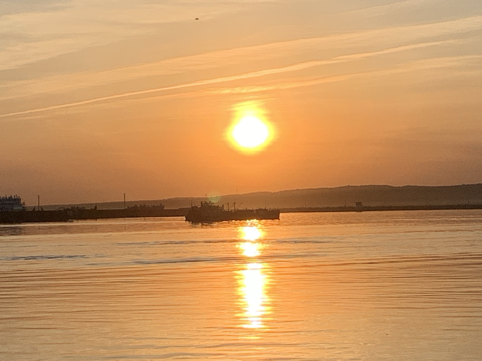 The image shows a sunset over water, likely the Kama River in the city of Chaikovsky, Russia. The sun is positioned high in the sky, reflecting brightly on the water's surface. In the distance, there's a shoreline with buildings and industrial structures. A few ships or barges are visible on the water near the shoreline. The sky has a warm, orange-yellow color gradient, and a few clouds are present with contrails.