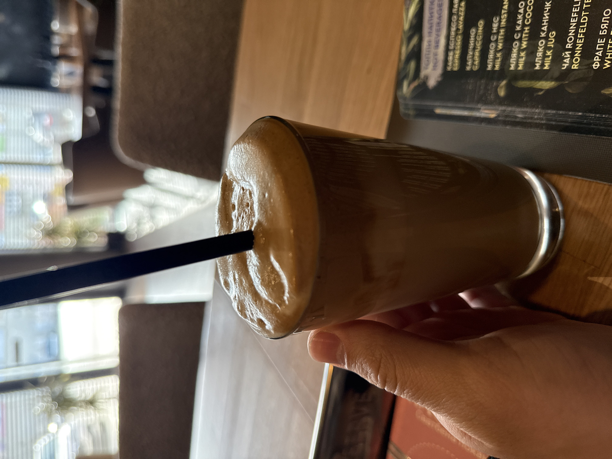 A close-up shot depicts a hand holding a glass of a foamy brown beverage, possibly iced coffee, with a black straw inserted. The glass sits on a wooden table next to a menu with text in Bulgarian and English, indicating items such as "Espresso Lavazza," "Cappuccino," "Milk with Instant," "Milk with Cocoa," "Milk Jug," and "Ronnefeldt Tea." The blurred background suggests an indoor cafe setting, identifiable by the menu as likely located in Sofia, Bulgaria.