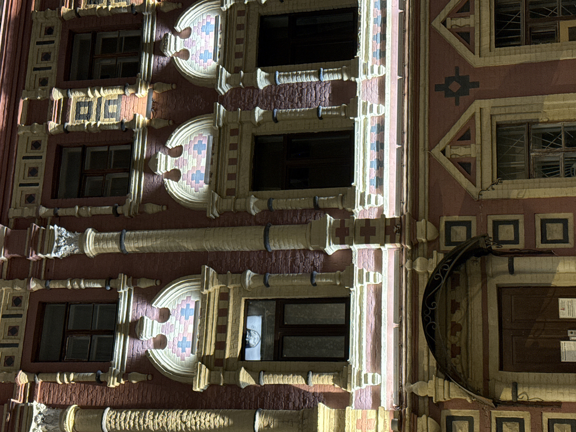 The image shows the facade of a historic building at night, illuminated by artificial lighting. The building is constructed of red brick and features ornate architectural details, including decorative columns, arched windows with patterned brickwork, and a portico above a set of double doors. The lighting highlights the textures of the brick and stone elements, creating shadows and contrasts. The windows are dark, suggesting they are either unlit or the interior is not visible. There are no people visible in the image. The overall impression is one of grandeur and historical significance. This building is identified as the Moscow State Literary Museum, located at Petrovka Street, 28, in Moscow, Russia.