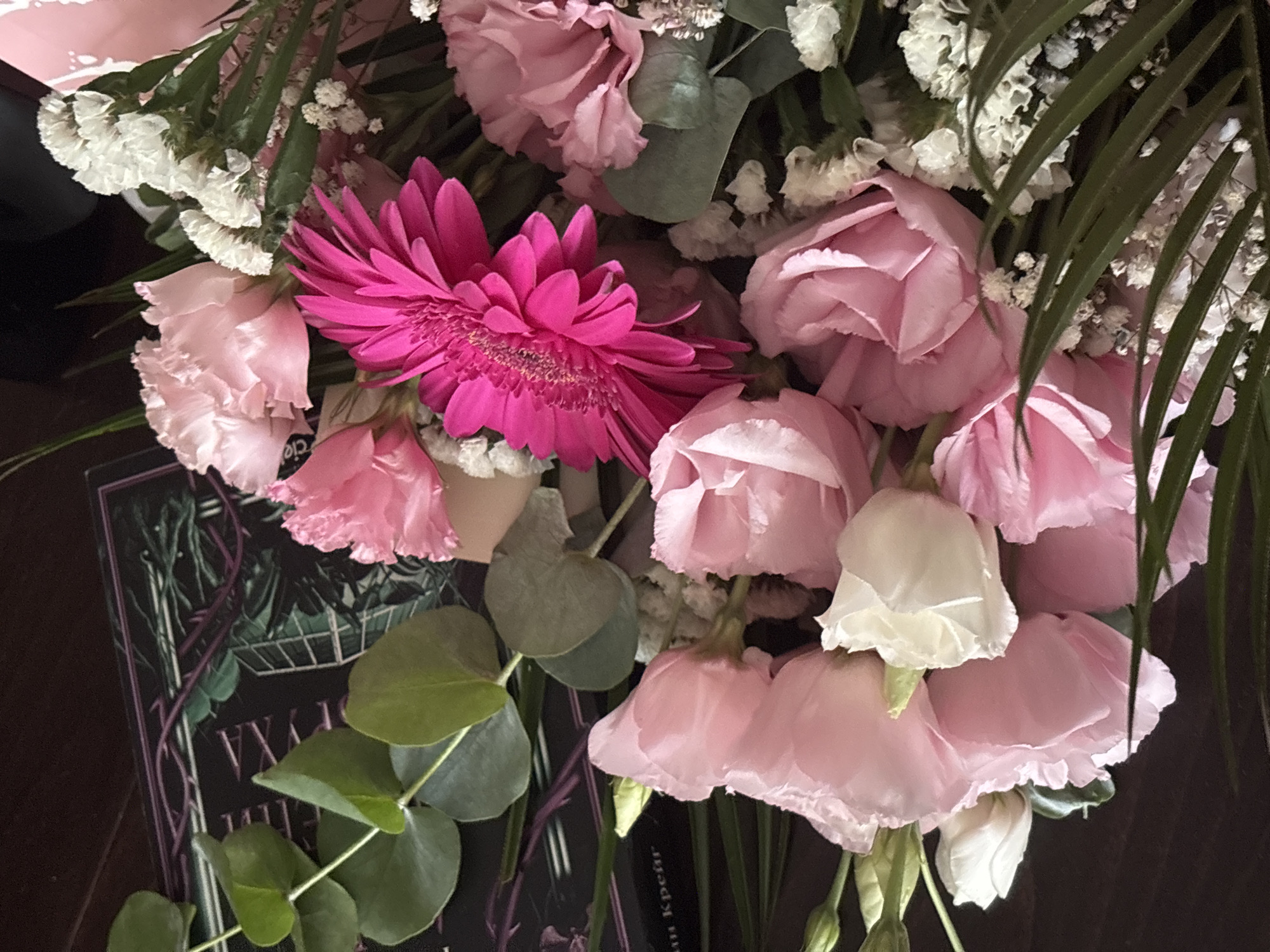 An overhead close-up shot shows a bouquet of pink and white flowers, along with eucalyptus leaves, resting on a dark wooden surface. The bouquet contains several pink roses, a vibrant fuchsia gerbera daisy, and clusters of small white flowers that appear to be baby's breath or statice. Amongst the flowers, several rounded, pale green eucalyptus leaves are visible, some of which are partially obscuring a dark book with a botanical illustration and purple vine-like designs on its cover. The book also has some text visible on its spine, though it is largely obscured by the flowers. The lighting suggests an indoor setting, possibly on a table or desk. There is no indication of the location being in Veliko Tarnovo, Bulgaria.