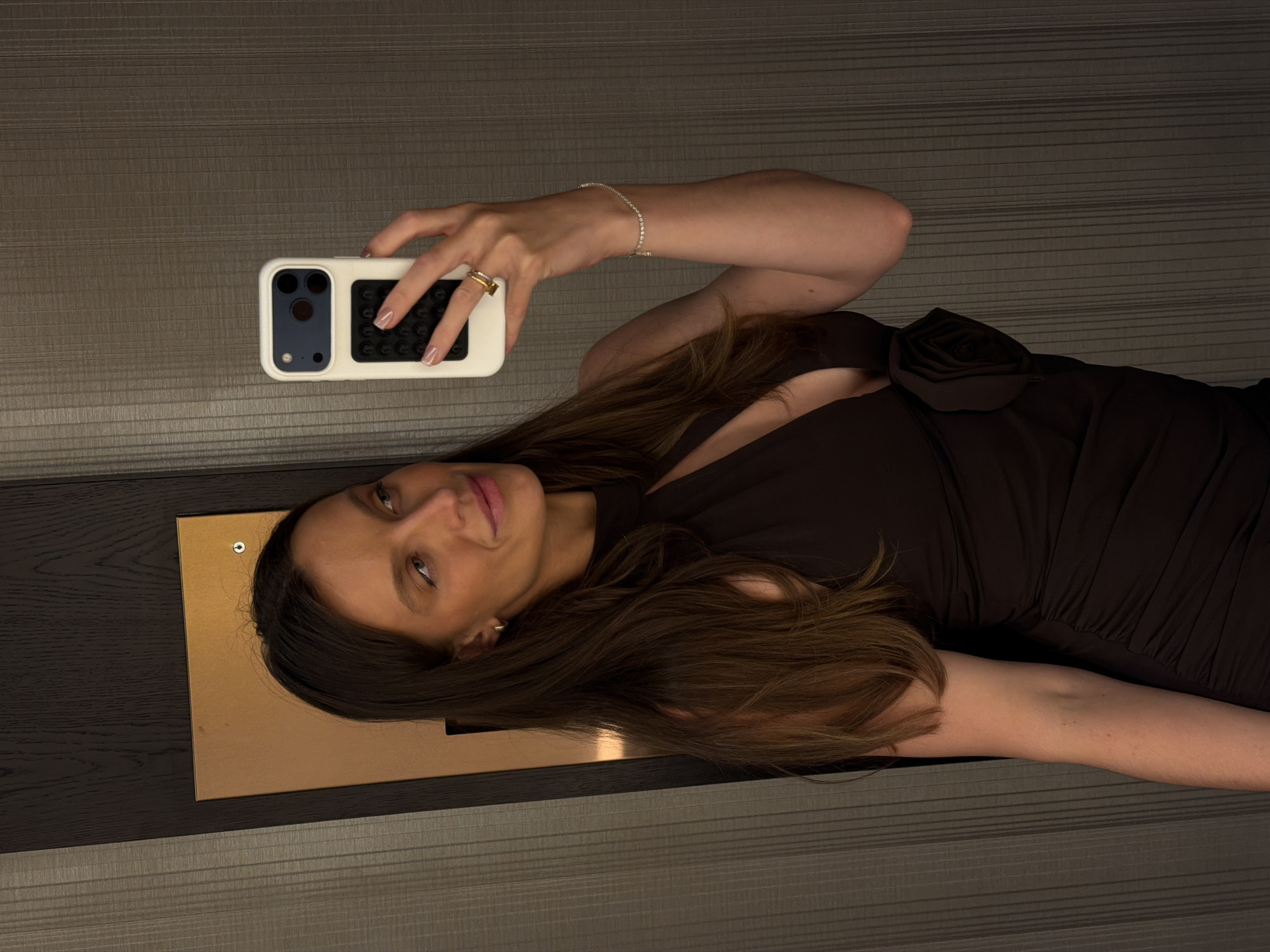 The image displays a woman taking a mirror selfie indoors, in Budva, Montenegro. She has long, dark brown hair and a tanned complexion, with her gaze directed slightly to her right and a subtle smile on her face. She is wearing a dark, possibly black or deep brown, sleeveless dress with a high halter-style neckline that includes a keyhole cutout on the chest. A prominent, fabric rose embellishment is attached to the left side of her dress, near her hip.
Her right hand holds a white smartphone, which features a dark blue/black camera module area and a distinct black rectangular grip on the back. Her fingers, adorned with light-colored nail polish in a French manicure style, are visible holding the device. A gold-toned ring with multiple bands is worn on her right index finger. On her left wrist, a thin, silver-colored chain bracelet with small, evenly spaced clear stones is visible. The background consists primarily of vertically textured grey-brown wall paneling. To the left and slightly behind the woman, a darker vertical wooden beam or frame is present, adjacent to a rectangular, copper-toned panel. A small, circular silver stud or screw is visible in the upper right corner of this copper panel.