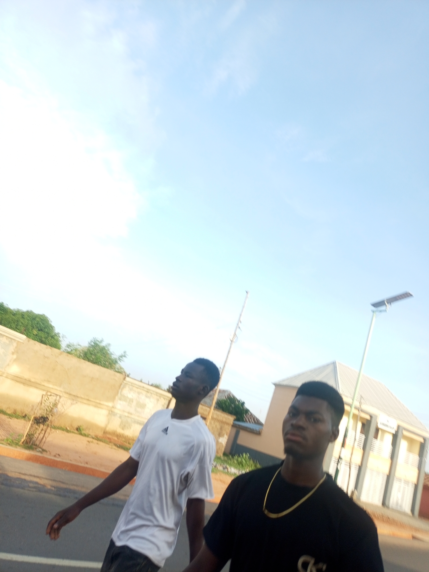 The image displays an outdoor daytime scene featuring two young adult males walking on a paved surface, likely a road or pathway. The sky occupies a significant portion of the upper frame, appearing clear and light blue with very sparse, wispy white clouds.
The subject positioned on the left, slightly further back in the frame, is a male with dark skin and short, dark hair. He wears a white short-sleeved t-shirt with a dark Adidas logo on the left chest and dark trousers or shorts. His head is tilted upwards and slightly to his right, and his gaze is directed off-frame, suggesting he is looking at something in the distance or above. His left arm is visible, hanging by his side.
The subject in the foreground on the right is also a male with dark skin and short, dark, neatly styled hair. He wears a black short-sleeved t-shirt, with a faint, lighter-colored logo (possibly "CK") visible on the lower left front. A prominent gold-colored, flat-link chain necklace adorns his neck. He is facing directly towards the viewer/camera, with a neutral to slightly serious expression and a direct gaze.
In the background on the left, a low, light brown or tan-colored wall or building facade is visible, topped with green foliage and trees. On the right side of the background, a two-story building with a light-colored exterior and a grey, pitched roof is present. Two vertical poles are also visible: one thin, light-colored pole to the left of the right subject, and a taller, light green pole on the far right, which features a solar photovoltaic panel mounted at its top, indicating a solar-powered street light. The ground beneath the individuals appears to be asphalt, transitioning to a reddish-brown dirt or gravel verge near the background structures. The lighting is bright and even, consistent with daylight.