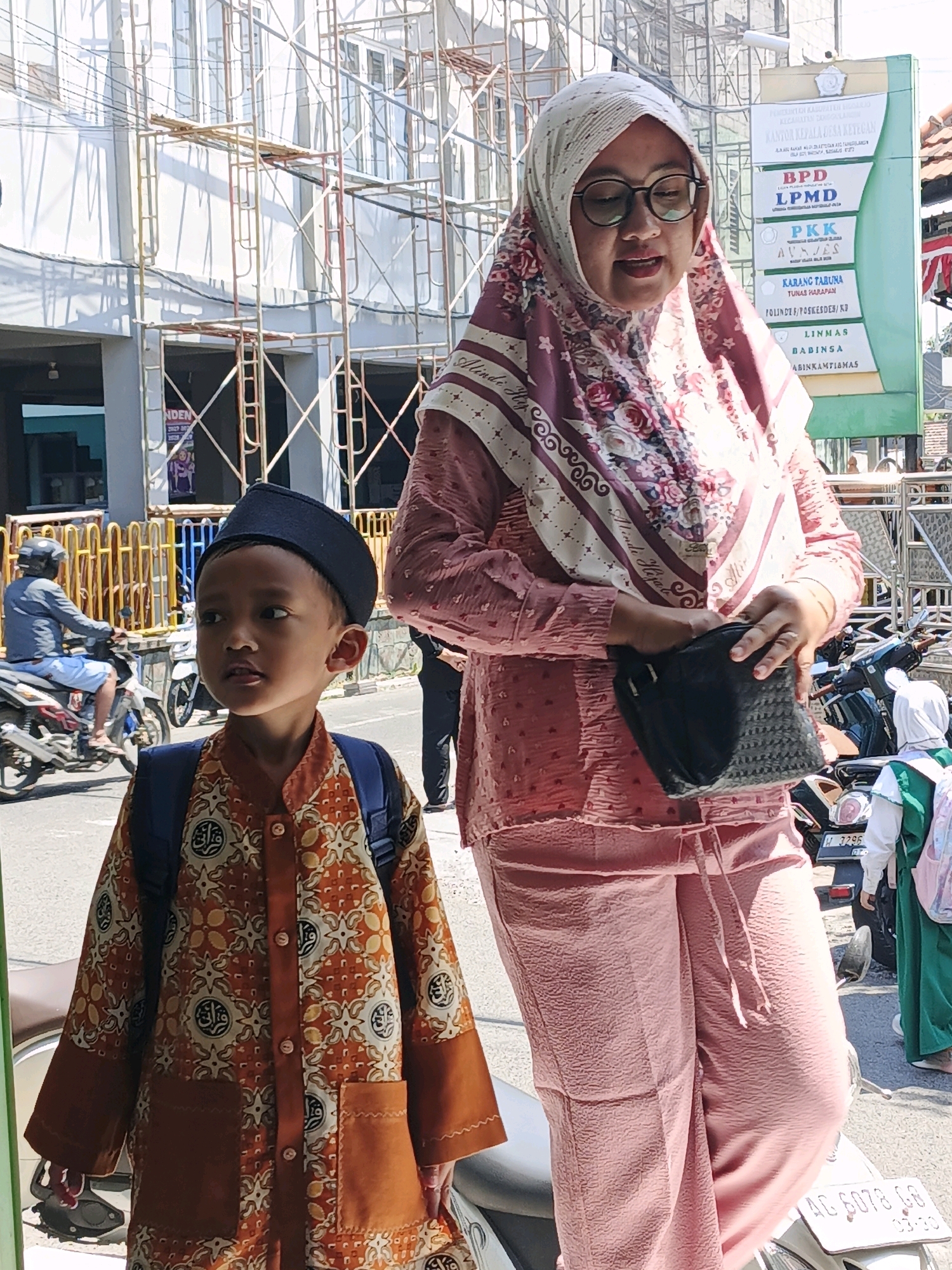 In the city of Ketegan, Indonesia, a boy wearing a black peci and a batik shirt stands on a street. To his right, a woman in a pink hijab and glasses holds a black purse. Behind them, scaffolding covers a building. A motorcycle with a rider is visible to the left, and other people and motorcycles are in the background. A sign board with text in Indonesian is also visible. The scene appears to be outdoors on a sunny day.