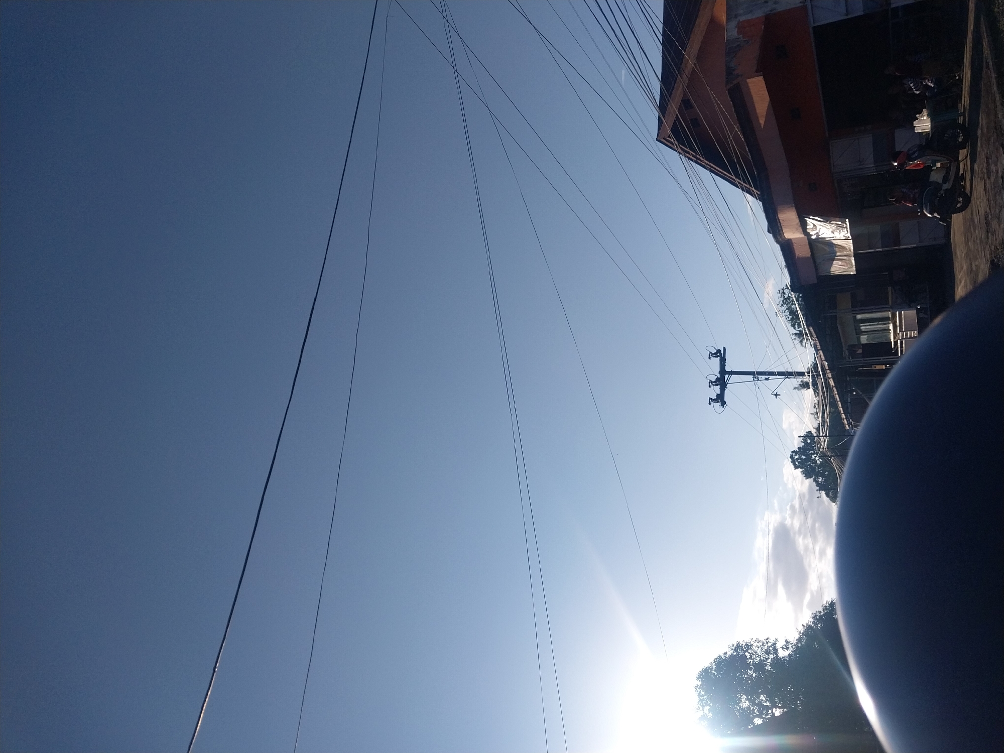 In Pallawa Rukka, Indonesia, a street scene unfolds under a clear blue sky with prominent power lines crisscrossing the frame. The sun, low on the horizon, casts a bright glare on the left side of the image, illuminating trees and some clouds. In the foreground, a dark, curved object, likely part of a vehicle's body, dominates the bottom of the frame. Beyond this, a street is visible, lined with buildings. On the right, a two-story building with an orange facade and a darker roof stands out. Several people are gathered in front of the building, near a scooter. A utility pole with electrical equipment is also present on the right side of the street.