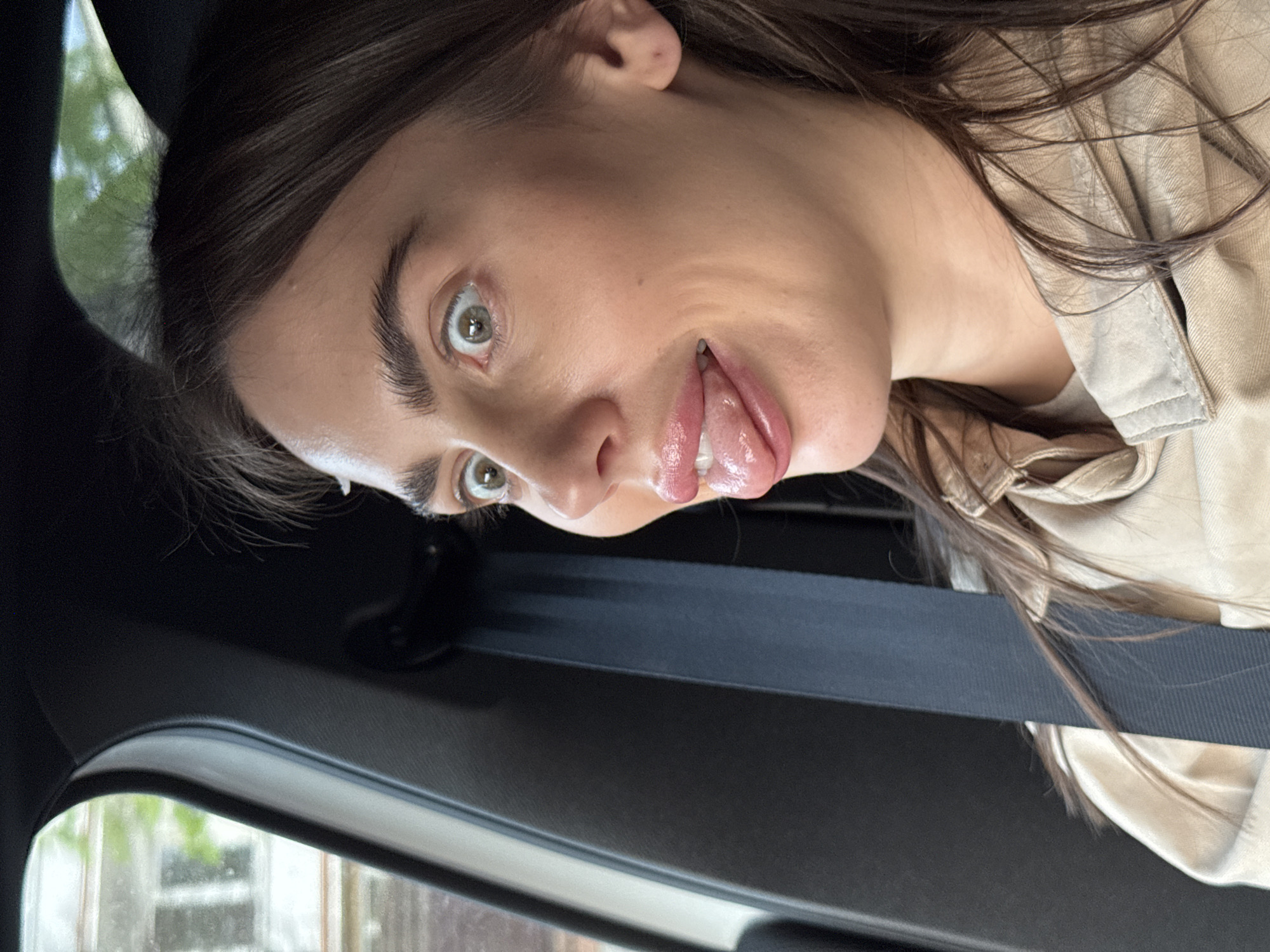 A woman with fair skin and dark brown hair is sticking her tongue out in a vehicle. She has light-colored eyes and is wearing a light brown jacket. The safety belt is across her chest. The window reveals an ambiguous outdoor background that does not provide identifiable elements for Warsaw, Poland.