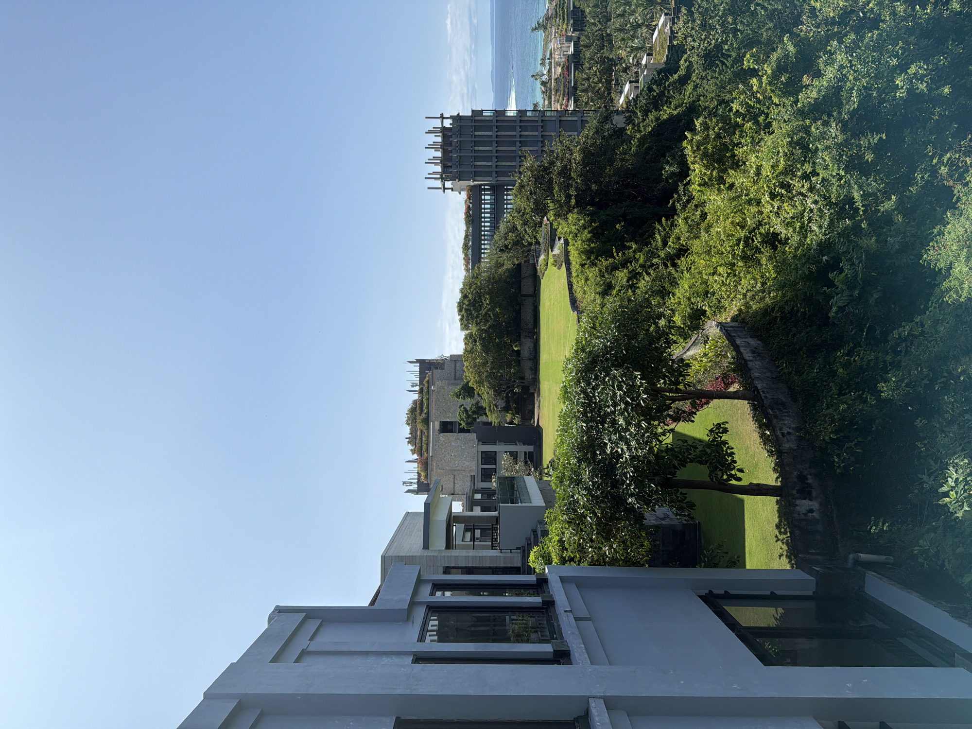 The image depicts a daytime view of Kuta Selatan, Indonesia. In the foreground, sections of modern, multi-level buildings painted in shades of gray are visible. These buildings feature large windows and minimalist architectural details. Beyond these, a neatly maintained grassy lawn stretches out, bordered by trees and a low stone wall. Further in the background, what appears to be a building under construction, characterized by an exposed concrete frame, stands prominently. In the distance, the sea meets the horizon, with an island visible along the coastline. The sky is a clear, light blue. Lush, dense foliage is also evident throughout the scene, interspersed with the buildings.