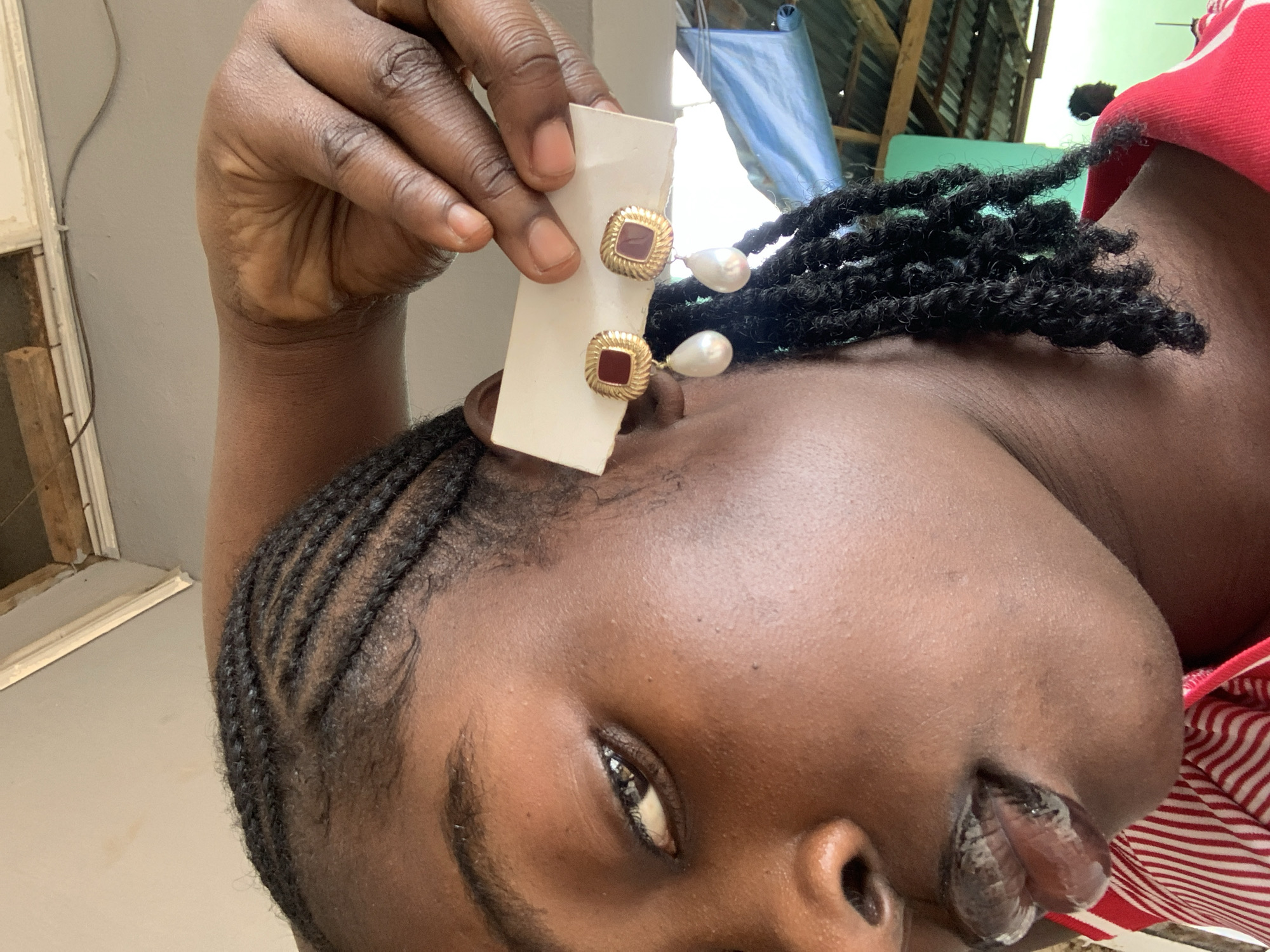 A close-up photograph captures a person with dark skin, shown from the chest upwards, their head tilted towards the right. Their left hand is visible, holding a small white card that displays two earrings. Each earring features a square, gold-toned frame with a ridged texture, enclosing a reddish-maroon central square. A white, teardrop-shaped pearl dangles from the bottom of each square. One earring is positioned very close to the person's earlobe, suggesting it is being displayed or tried on.
The person has distinct hairstyles: cornrows are visible on the side of their head, neatly braided, leading into a section of dark, twisted or dreadlocked hair that rests on their shoulder. A garment with red and white stripes is seen on their upper body. The person's visible eye, dark and with a clear reflection, looks directly at the viewer, and their mouth is slightly ajar, revealing part of their tongue.
The background is an informal mix of elements. To the left, a light-colored wall with a wooden frame or window structure is visible. The upper right background reveals components of an outdoor or semi-outdoor setting, including a light blue tarp, a green surface, and exposed wooden planks, suggestive of a basic structure or roofing. The natural, bright lighting indicates it is daytime. This scene is contextualized in New Stadium, Nigeria. No visible text is present in the image.