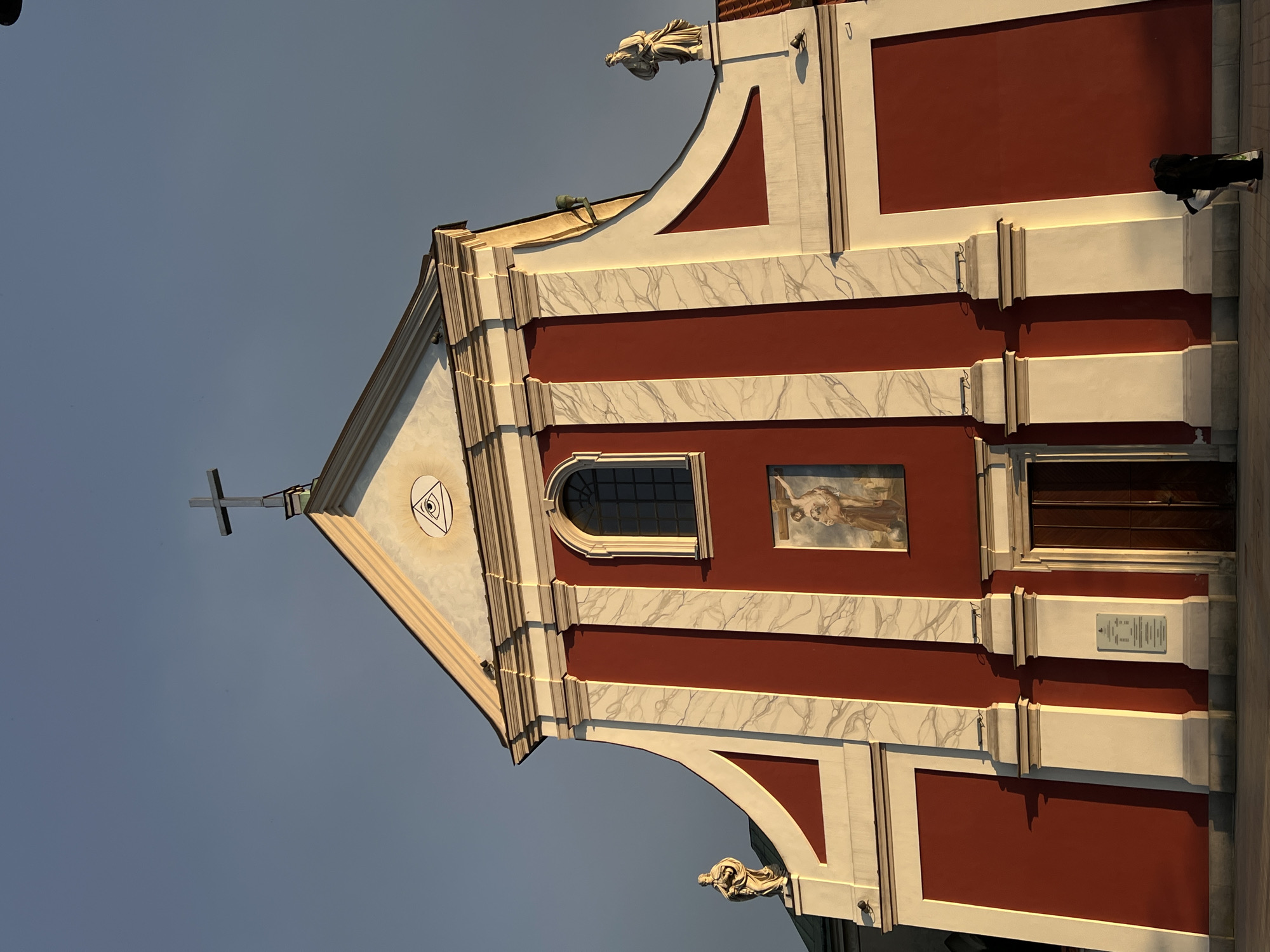 The image shows the facade of a church in Lublin, Poland. The church has a red and white exterior with a large cross on top. There are two statues on either side of the facade, and a painting of Jesus Christ above the entrance. The sky is clear and blue. A person is walking away from the church in the foreground.