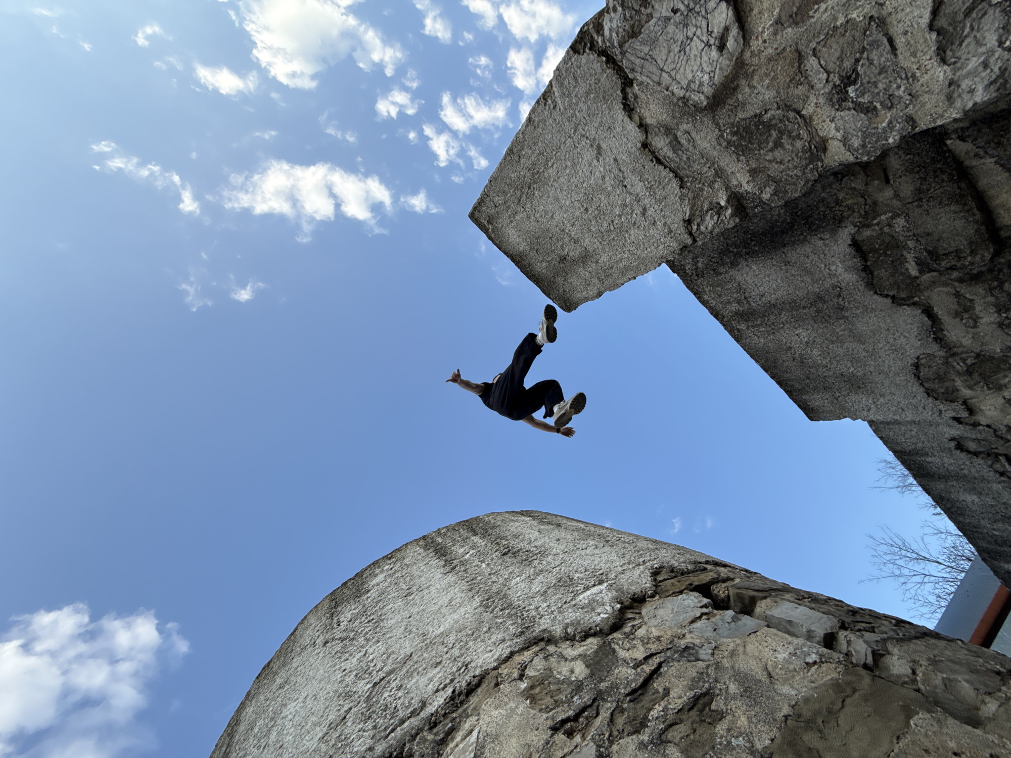 A low-angle, color photograph shows a person jumping between two large, gray, stone structures against a blue sky with scattered clouds. The person is in mid-air, wearing dark clothing and light-colored shoes, with arms extended. The stone structures appear to be weathered and aged. The location is Veliki Preslav, Bulgaria.