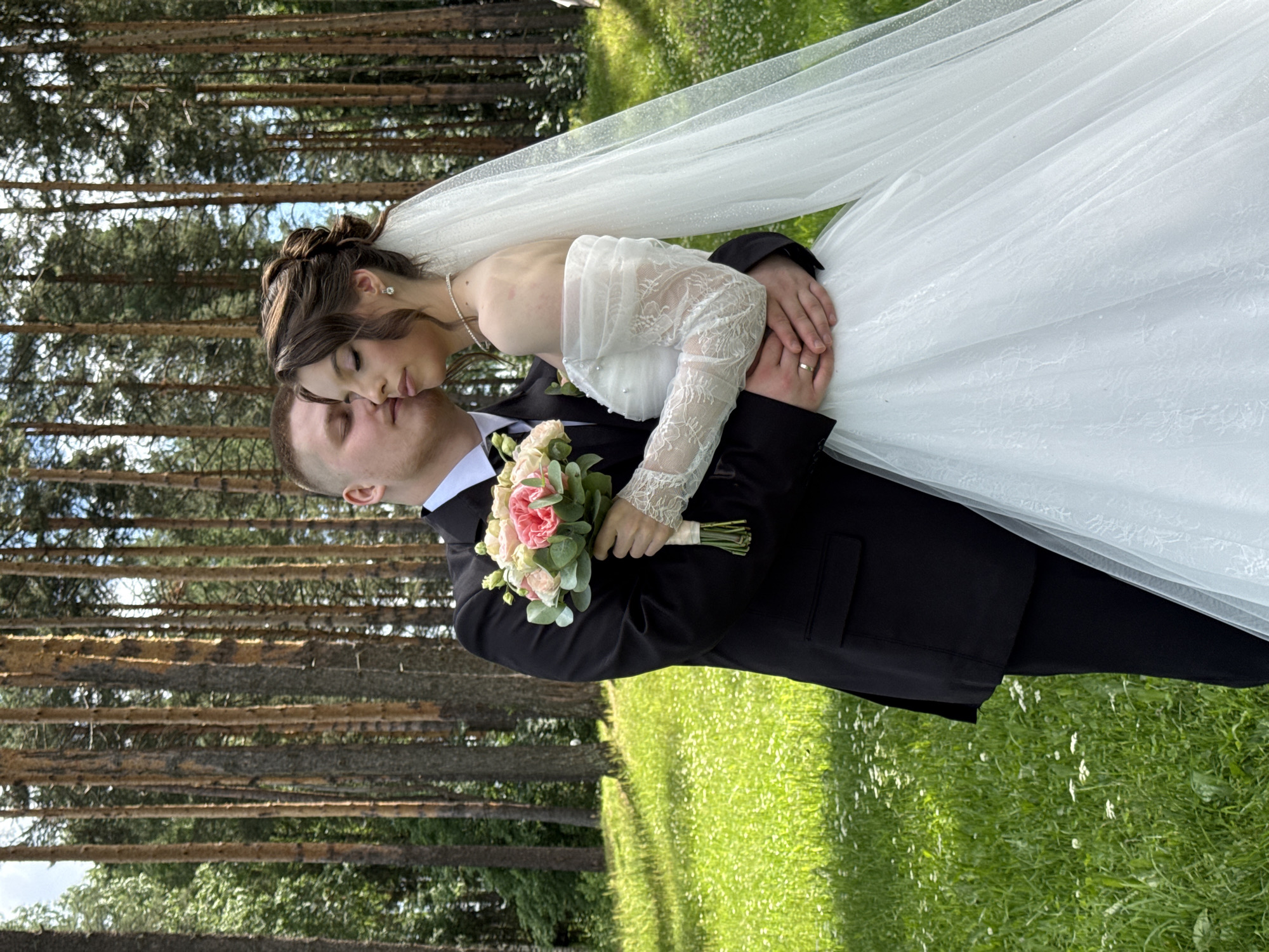 A wedding couple is posing for a photo in a forest clearing with tall pine trees in the background. The groom, wearing a black suit and white shirt, is embracing the bride, who is in a white wedding dress with lace sleeves and a veil. She is holding a bouquet of pink and white flowers. The ground is covered in green grass with small white flowers. The scene appears to be in the city of Votkinsk, Russia.