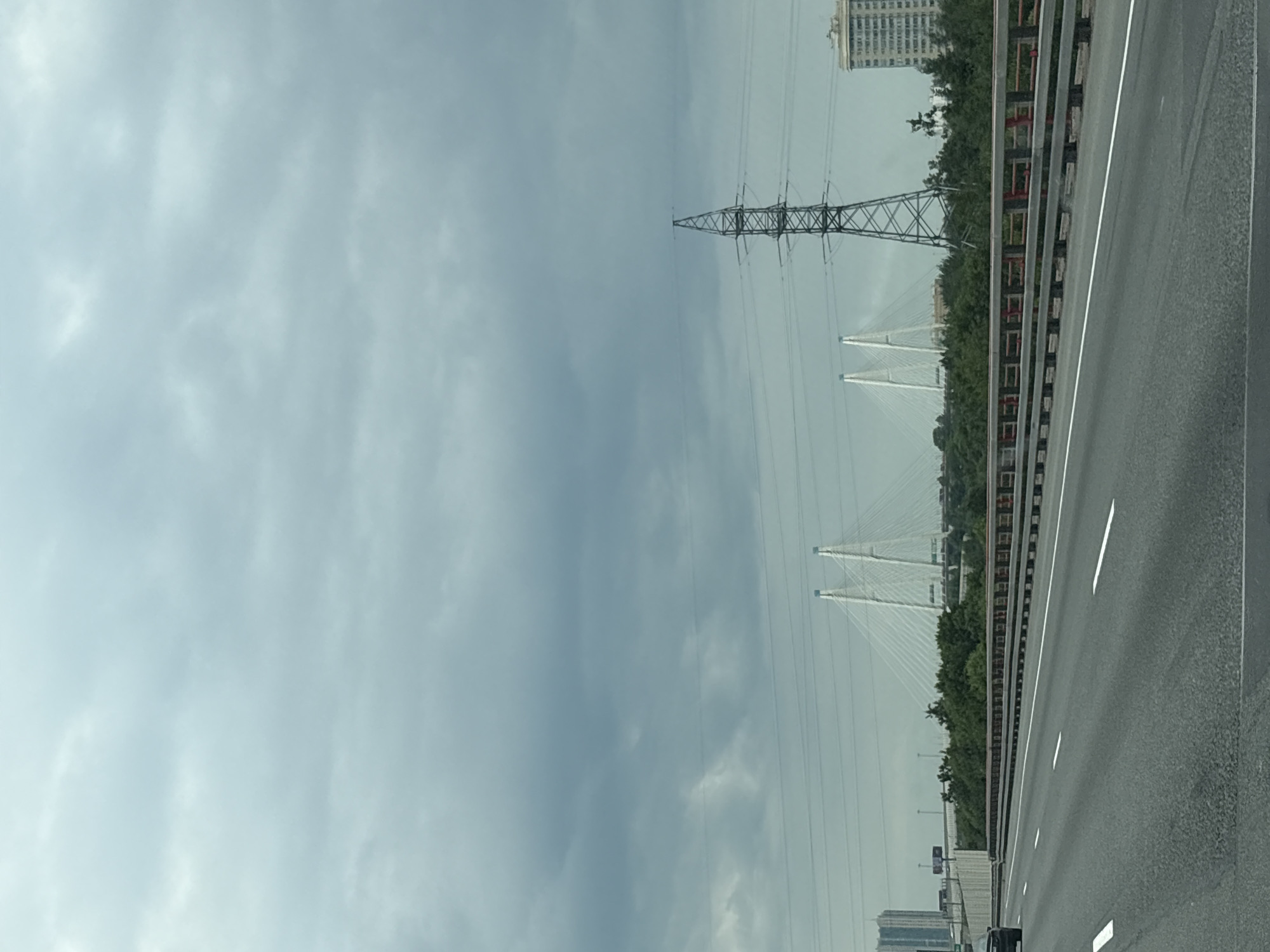 The image shows a highway in Novosaratovka, Russia, on a cloudy day. The highway is paved with asphalt and has white lane markings. A concrete barrier with red and yellow accents separates the highway from the median. On the median, there are trees and shrubs. In the background, there is a large cable-stayed bridge with white towers, an electricity pylon, and a tall building. A car is visible driving on the highway. The sky is overcast with gray clouds.