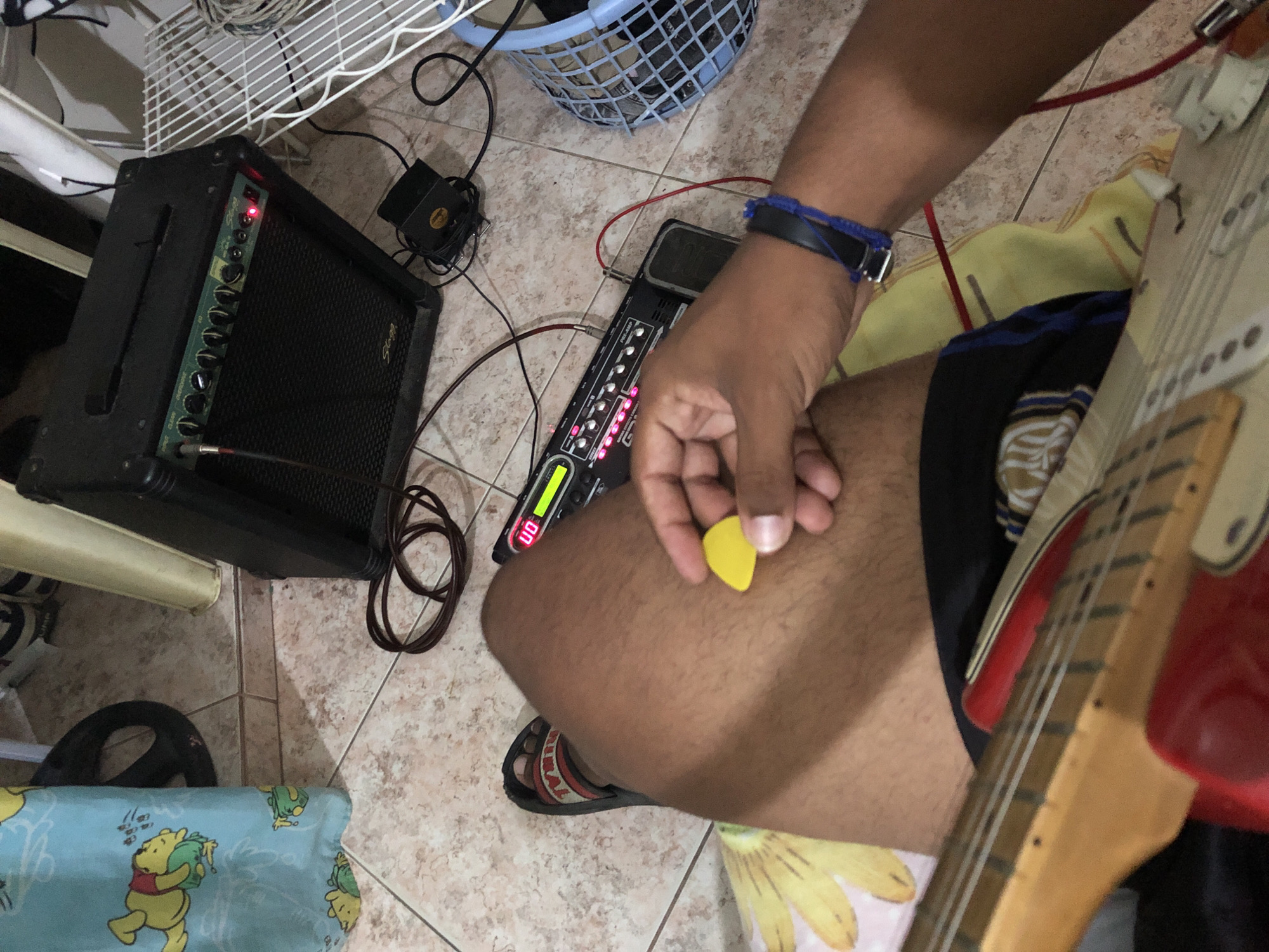 A person is seated, holding a yellow guitar pick in their right hand, with a red electric guitar visible on the right. To the left are a small black guitar amplifier and a multi-effects pedalboard connected by cables. The floor is tiled, and various household items, including a laundry basket, a fan, and furniture, are present. The location is implied to be Altagracia, Venezuela.