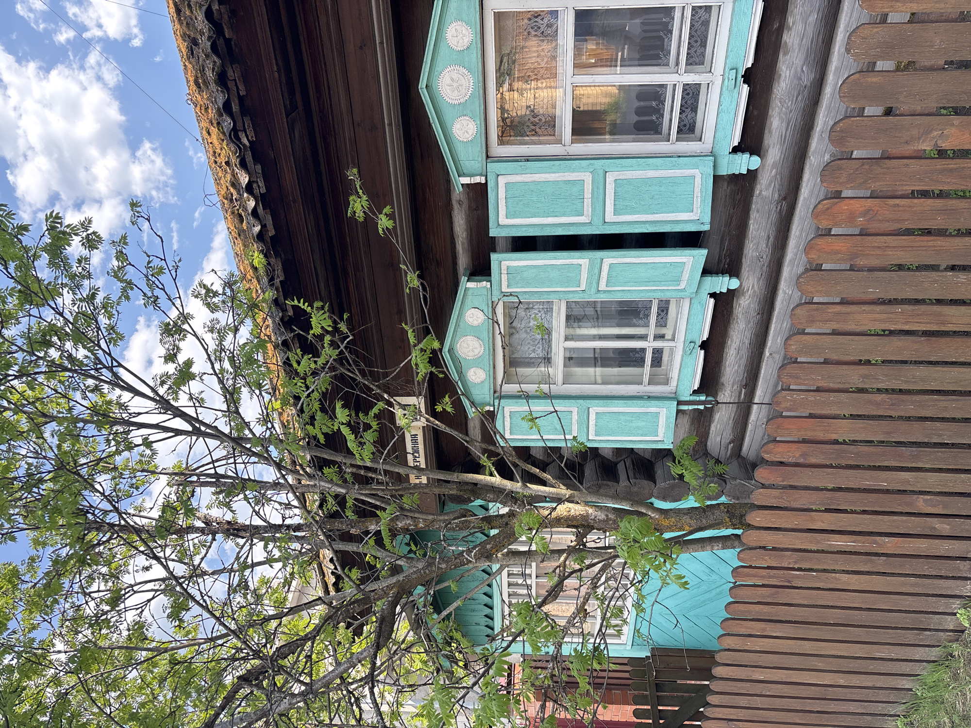 The image depicts a traditional wooden house in the city of Овсянка, Russia, partially obscured by a tree. The house features two windows with ornate turquoise and white painted frames. A brown wooden fence stands adjacent to the house. The sky is visible through the tree branches, with scattered clouds.