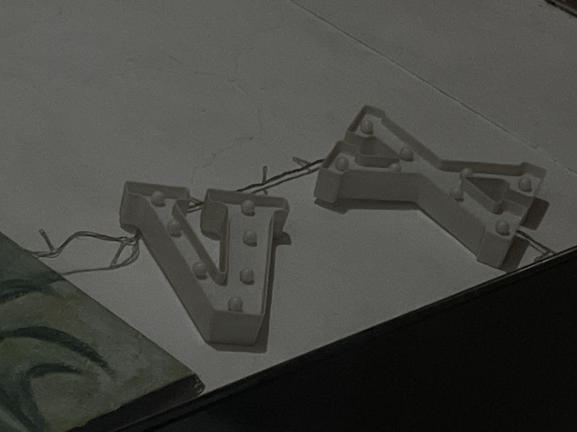 The image depicts a dimly lit indoor scene in Santa Ana, Venezuela, focusing on two unlit decorative marquee-style letters arranged vertically on a light gray, textured surface. The upper object is a white number '4', featuring several unlit, circular bulbs embedded along its frame. Below it, connected by thin, dark electrical wires, is a white letter 'Y', also equipped with multiple unlit, circular bulbs. Additional wires extend upwards from the '4' and downwards from the 'Y'. In the upper left quadrant, a portion of a painting or mural is visible, characterized by swirling patterns in dark green and lime green hues. The surrounding areas are dark and indistinct, suggesting shadows or the periphery of the illuminated space. No individuals or active interactions are present.